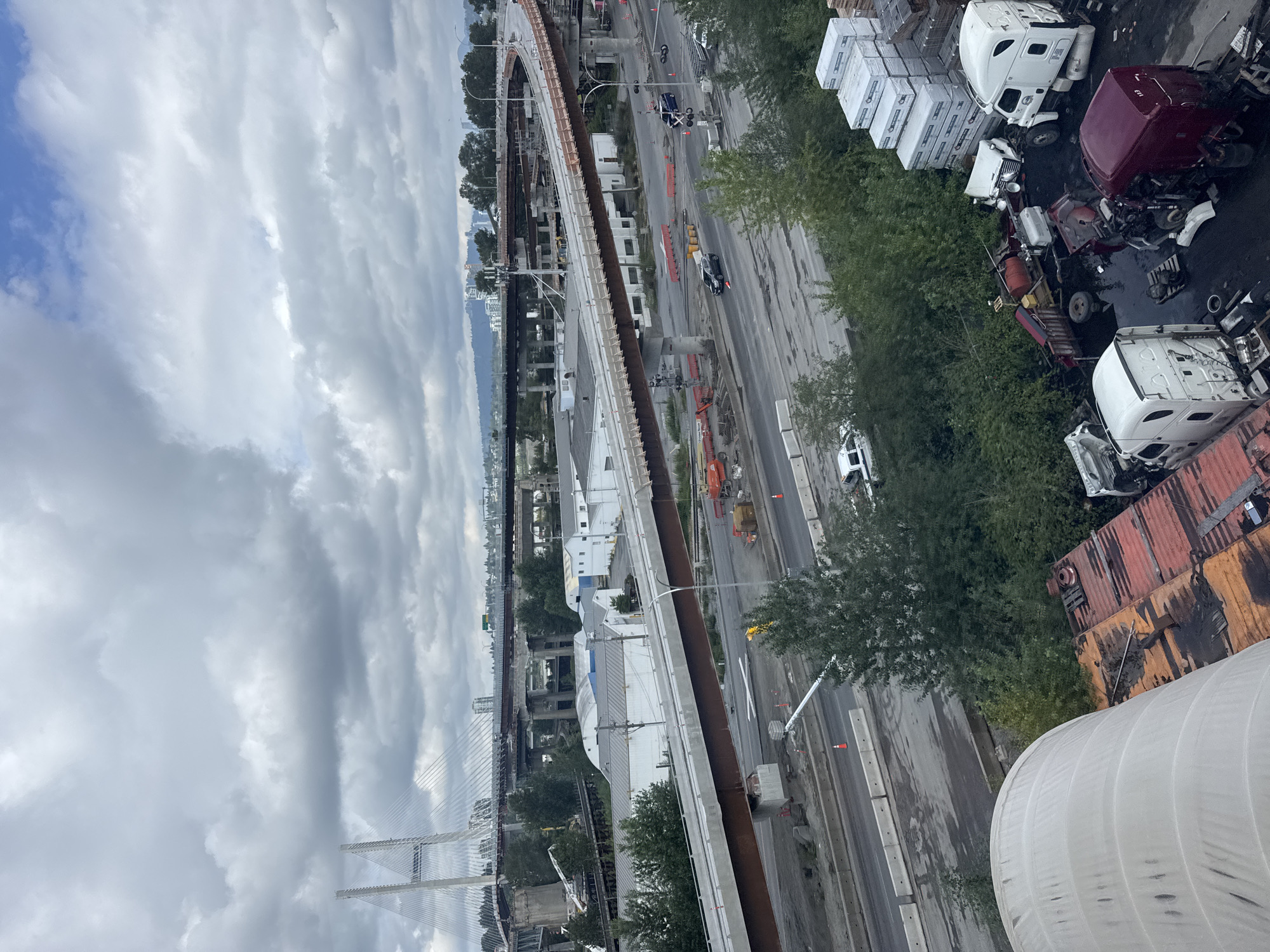 The image captures an aerial view of a busy urban construction site in Surrey, Canada, characterized by extensive infrastructure development, specifically focusing on a multi-level highway overpass. The overcast sky dominates the upper portion of the frame, with a large suspension bridge visible in the distance. Below, the complex network of the highway under construction is the focal point, with concrete barriers, orange construction cones, and various heavy machinery like cranes and excavators scattered throughout the site. Several vehicles, including a police car and other cars, are visible on the roadways, suggesting ongoing traffic flow amidst the construction. In the foreground, a collection of disassembled truck parts, including cabs and engines, are piled up, possibly indicating a repair or salvage yard. The scene is a mix of ongoing development and industrial activity, typical of a rapidly growing urban environment.