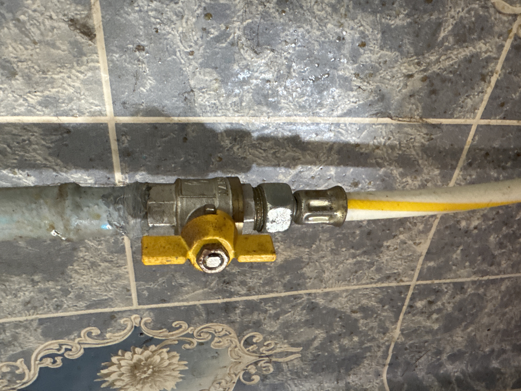The image shows a close-up of a gas valve and hose attached to a wall covered in blue tiled wallpaper. The gas valve is yellow and silver. A white and yellow hose is connected to the valve. The wallpaper has a decorative floral pattern on the left side. The image appears to be taken indoors. The specific location in Rozdilna, Ukraine, is not discernible from the image itself.