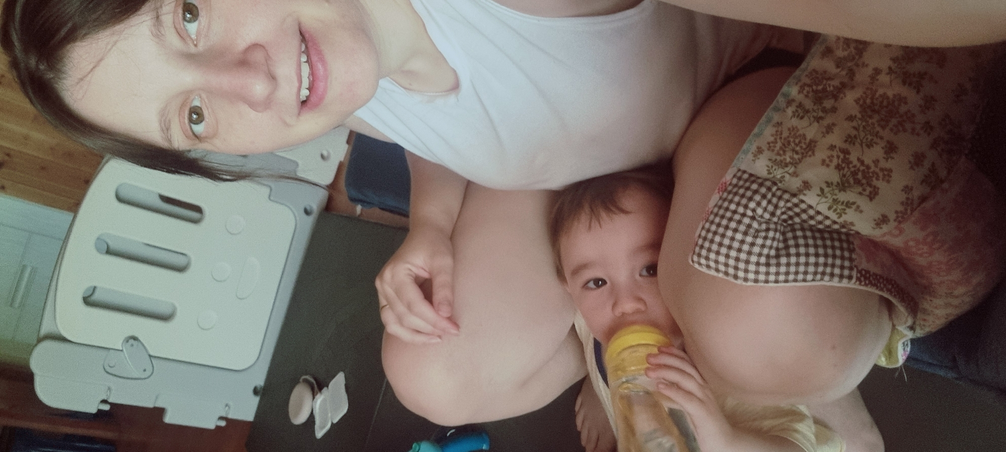 A woman and a young child are visible in an indoor setting. The woman is fair-skinned and wearing a white tank top. She appears to be taking a selfie. The child is fair-skinned and holding a bottle. A patchwork pillow is near them. A gray playpen is visible in the background, along with baby essentials. Based on image metadata, the location is Dunnae-myeon, South Korea.
