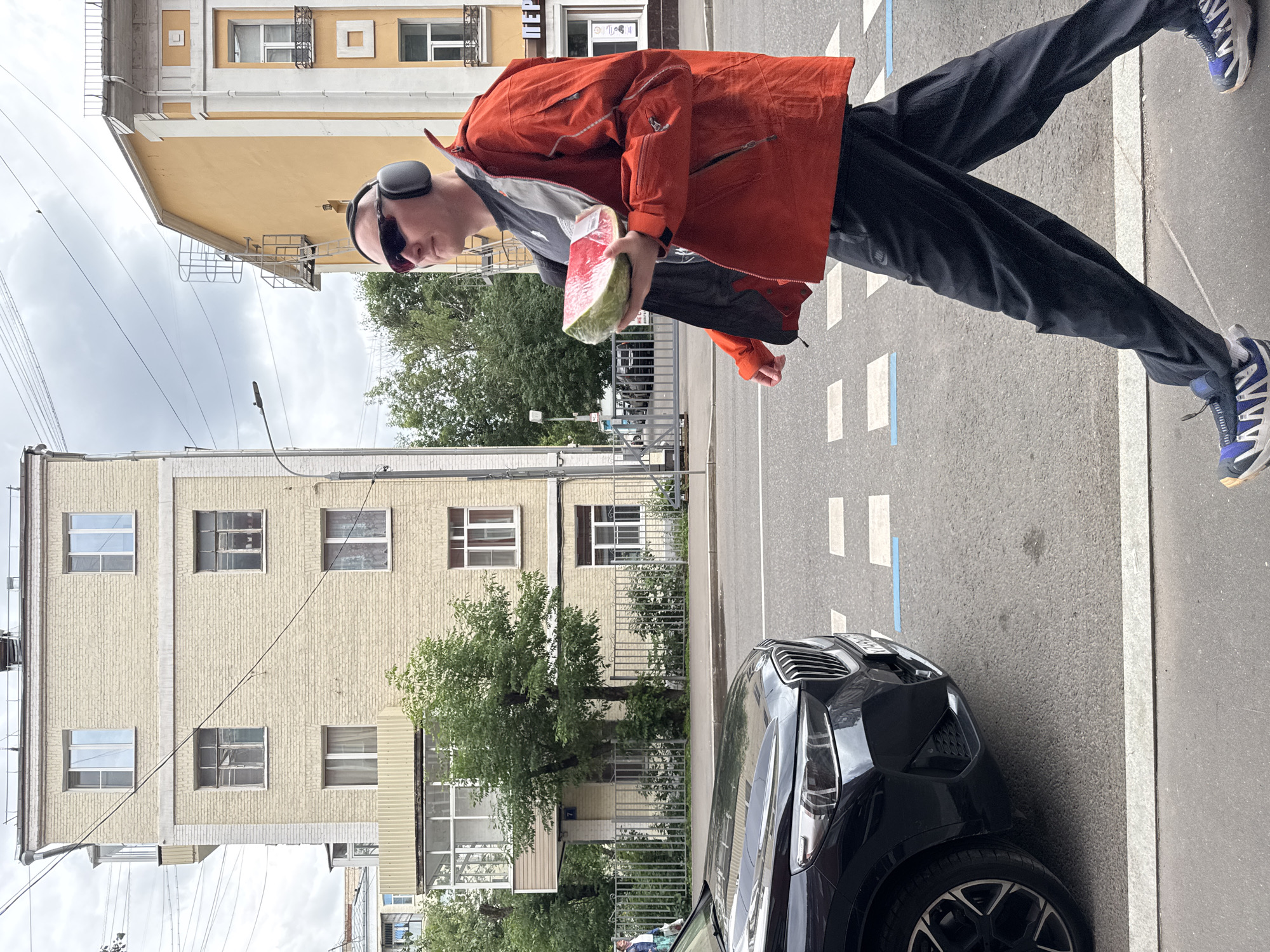 A man walks on a city sidewalk in Moscow, Russia, wearing headphones, sunglasses, an orange jacket, black pants, and athletic shoes. He is carrying a large slice of watermelon wrapped in plastic. In the foreground, a black car is parked on the street, and a crosswalk is visible. Behind the man, there are buildings, trees, and utility wires against a cloudy sky. The man is walking towards the right side of the frame.