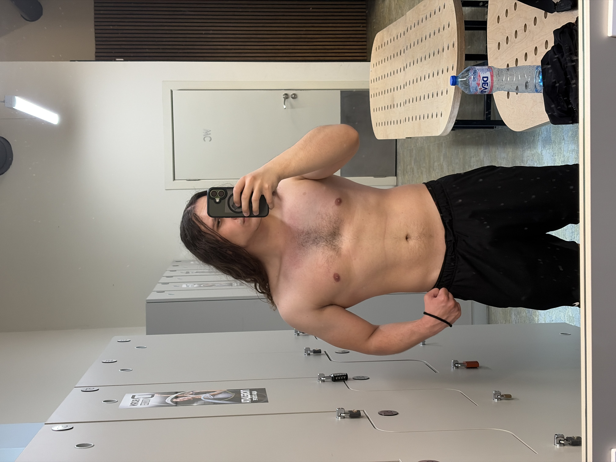 A young man is captured in a shirtless mirror selfie, likely within a locker room or gym changing area in Plovdiv, Bulgaria. He has long, dark hair that falls over his shoulders and is wearing black athletic shorts. His face is obscured by the smartphone he holds up to take the photo, which is encased in a dark protective cover.
The reflected background reveals key features of the facility. To the left, a white door is visible, marked with the letters "MC," possibly indicating a restroom or men's changing room. Above this, a long, horizontal fluorescent light fixture provides illumination. Further back, a section of wooden slatted wall paneling adds a textural element to the upper part of the room. To the right, a light-colored wooden bench or table with a distinctive perforated pattern holds a clear plastic bottle of "DEVIN" brand water, which is a common Bulgarian brand, consistent with the location context. A black bag or item of clothing rests beside the bottle.
In the foreground, a row of grey lockers stretches into the distance. Several lockers are secured with combination locks, while others feature traditional key locks. One locker displays a sticker with text partially visible as "АТЛЕТИЧЕСКА ЗАЛА," meaning "Athletic Hall" or "Gym" in Bulgarian, confirming the athletic facility setting. The floor appears to be a neutral-toned, possibly concrete or tiled surface. The bright, even lighting suggests an indoor public space, and the man's pose is a casual one for a personal photo.