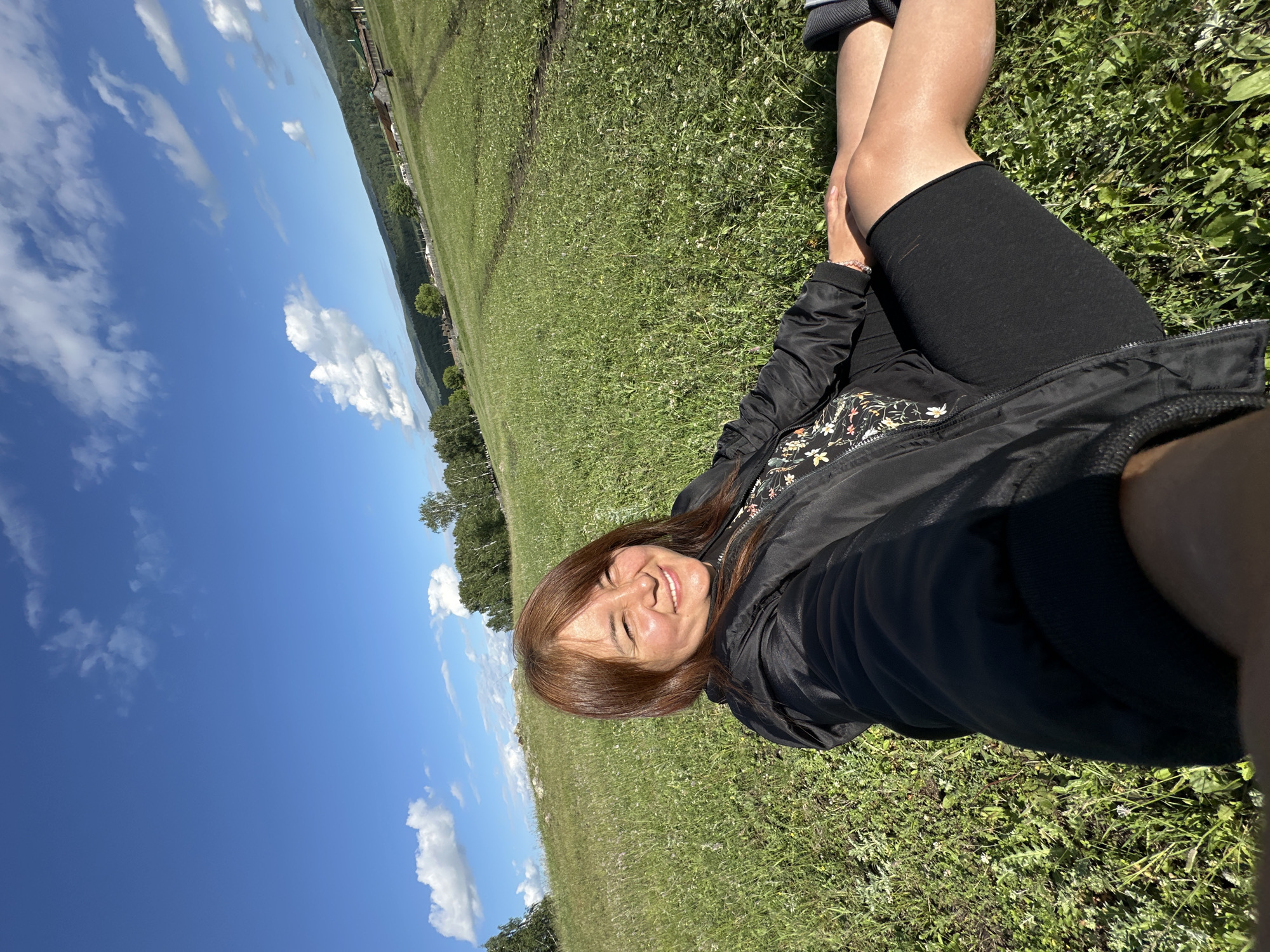 An adult female with light brown hair is lying on a grassy field, taking a selfie. She is wearing a black jacket over a floral top and black shorts. The field is green and covered with small white flowers. In the background, there are trees, hills, and a few houses. The sky is blue with white clouds.