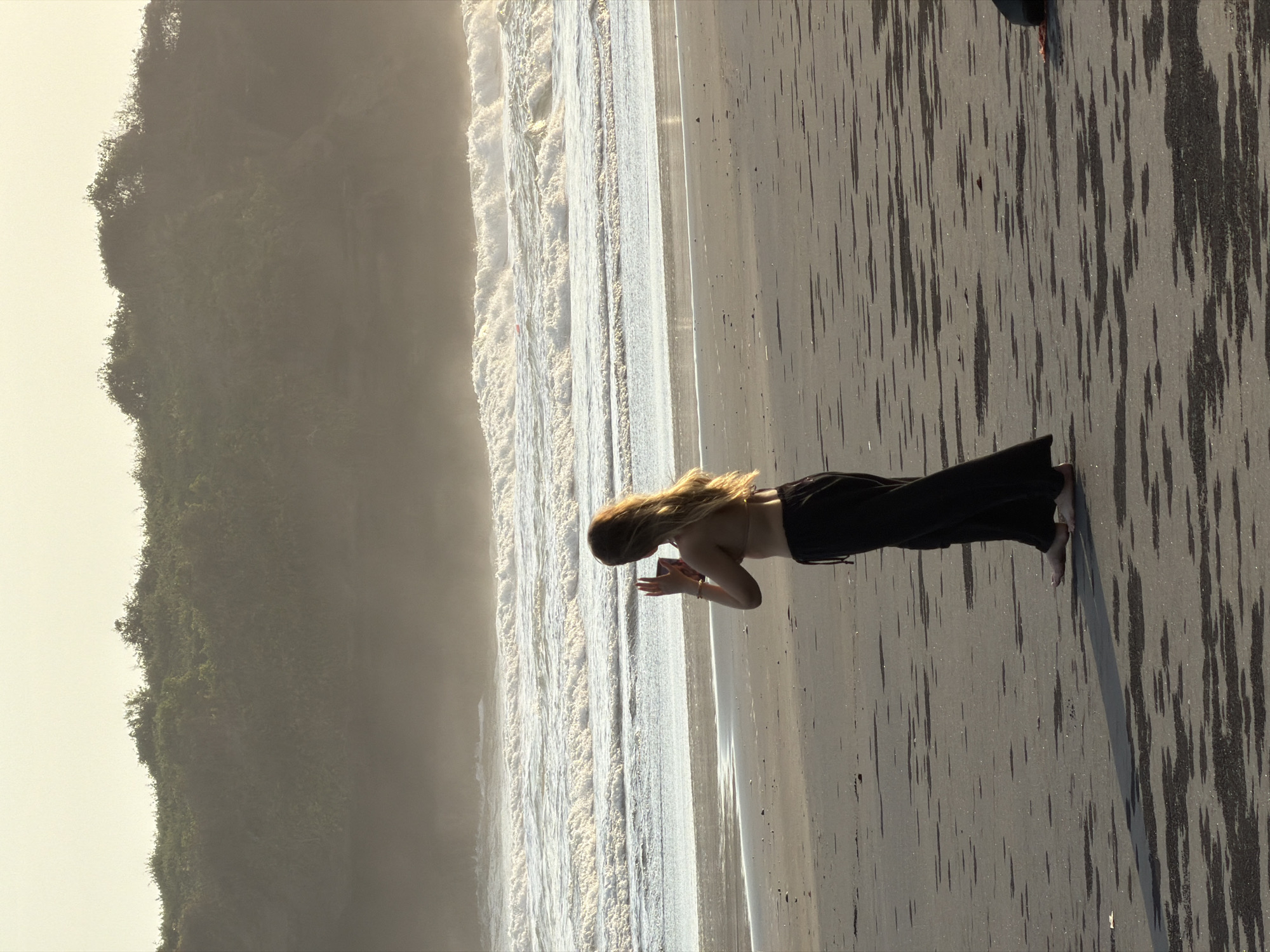 A full-shot, eye-level, medium-distance photograph captures a young woman with long blonde hair standing on a beach in Selemadeg, Indonesia. She is facing away from the camera, towards the ocean, and appears to be taking a photo with her phone. She is wearing a light-colored bikini top and dark, flowing pants. The beach is composed of dark sand with patterns of wet sand. In the background, large waves crash on the shore, and a lush, green cliffside rises behind the ocean. The sky is a pale yellow, suggesting either sunrise or sunset. A small, dark object, possibly a bag, is visible on the sand to the right of the woman.