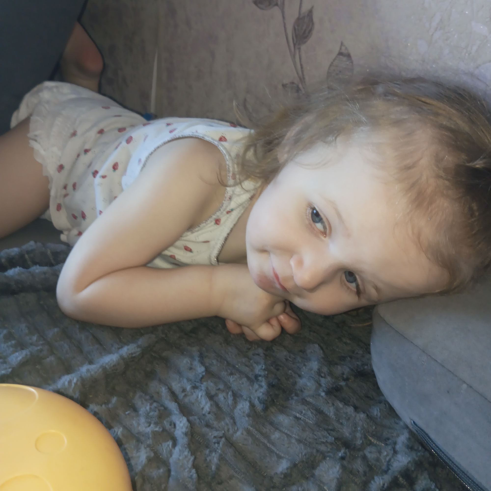 A young child, likely female, is lying on what appears to be a grey, textured blanket or rug. The child is resting against a light grey upholstered object, possibly a sofa arm or cushion. She is wearing a light-colored top with a small strawberry print. The child has fair skin, light brown hair with some curls, and blue eyes. Her hands are clasped together near her face. A portion of a yellow, round object with circular indentations is visible in the lower left corner of the frame. The background features a textured wall with a floral pattern. The location is Chaikovsky, Russia.