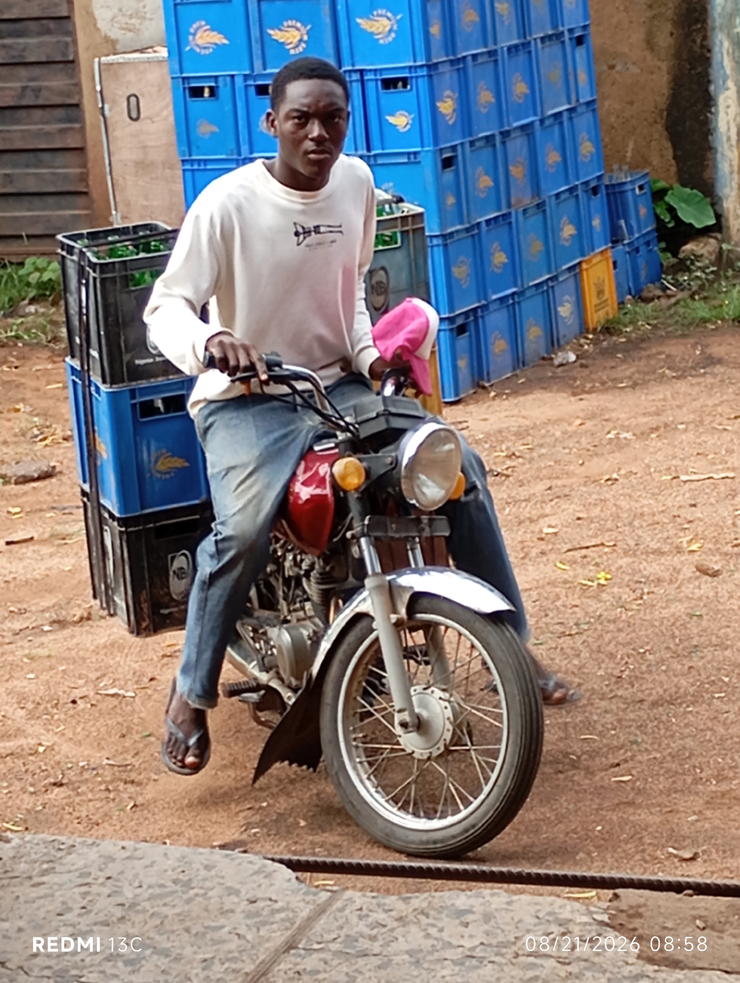 A young Black male, appearing to be in his late teens or early twenties, is seated on a red and silver motorcycle, facing forward and looking towards the camera. He is wearing a long-sleeved white shirt and blue jeans. The motorcycle has a prominent headlight and a spoked front wheel. Stacked behind the rider are several black and blue plastic crates, some appearing to contain green bottles. To the left of the motorcycle, a wooden structure with a visible number "0" is present. In the background, rows of blue plastic crates are stacked high, and a concrete wall is visible on the right. The ground is covered in reddish-brown dirt. The scene appears to be outdoors, likely on a street or in a yard within the city of Shendam, Nigeria. The timestamp in the bottom right corner indicates the image was taken on August 21, 2026, at 08:58.