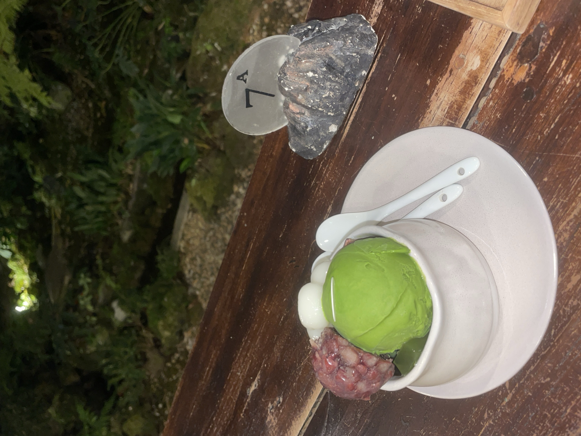 The image shows a wooden table with a plate of dessert, a table number, and a rock. The dessert consists of a scoop of green ice cream, small white dumplings, and red bean paste in a white bowl on a light pink plate. Two small white spoons are on the plate. A table number "7A" is visible next to a dark-colored rock. The table appears to be outdoors, with greenery in the background. The location is Chiang Mai City Municipality, Thailand.