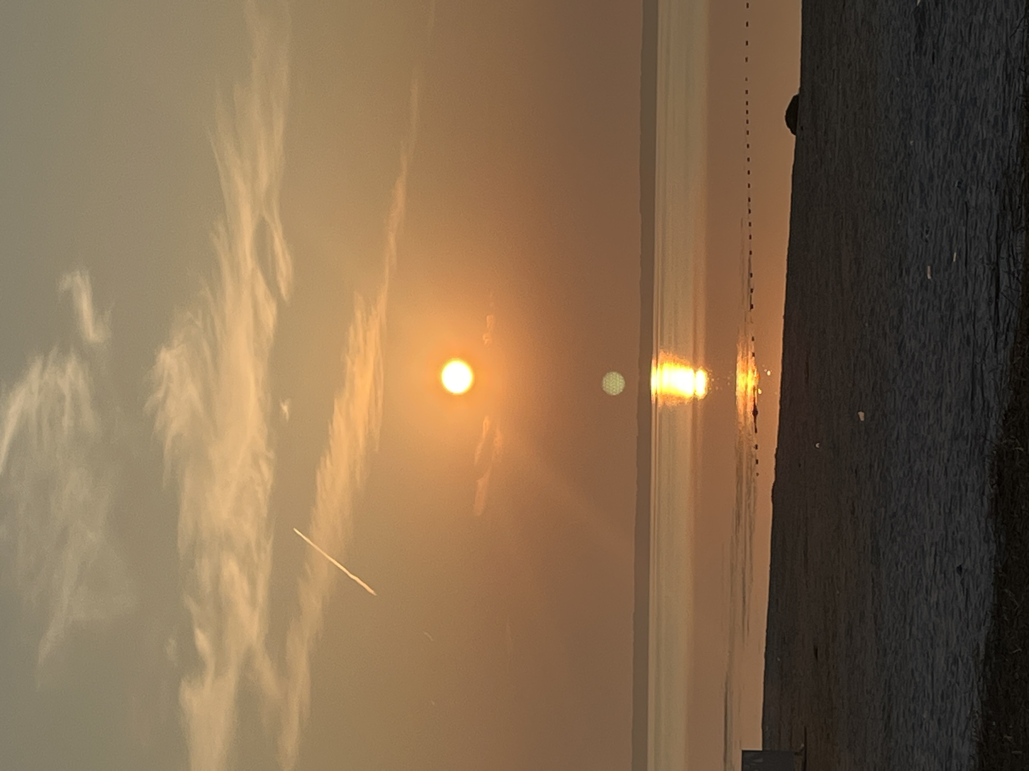 The image depicts a sunset over a body of water, likely a bay or sea, with a sandy beach in the foreground. The sky is filled with a warm, orange glow, characteristic of a sunset. wispy clouds are scattered across the sky, with a contrail from an airplane visible high up. The sun, a bright orange orb, is positioned in the middle of the frame, its reflection creating a shimmering path on the water. A faint, blurred light spot, possibly a lens flare or another reflection, is visible below the sun on the water. The water's surface appears calm and reflects the colors of the sky. The beach is a strip of sand, appearing dark in the low light conditions, with some scattered debris. In the far distance, a dark, horizontal line suggests the presence of a distant shoreline or landmass. The scene is tranquil and natural. The location is identified as Surakovo, Russia.
