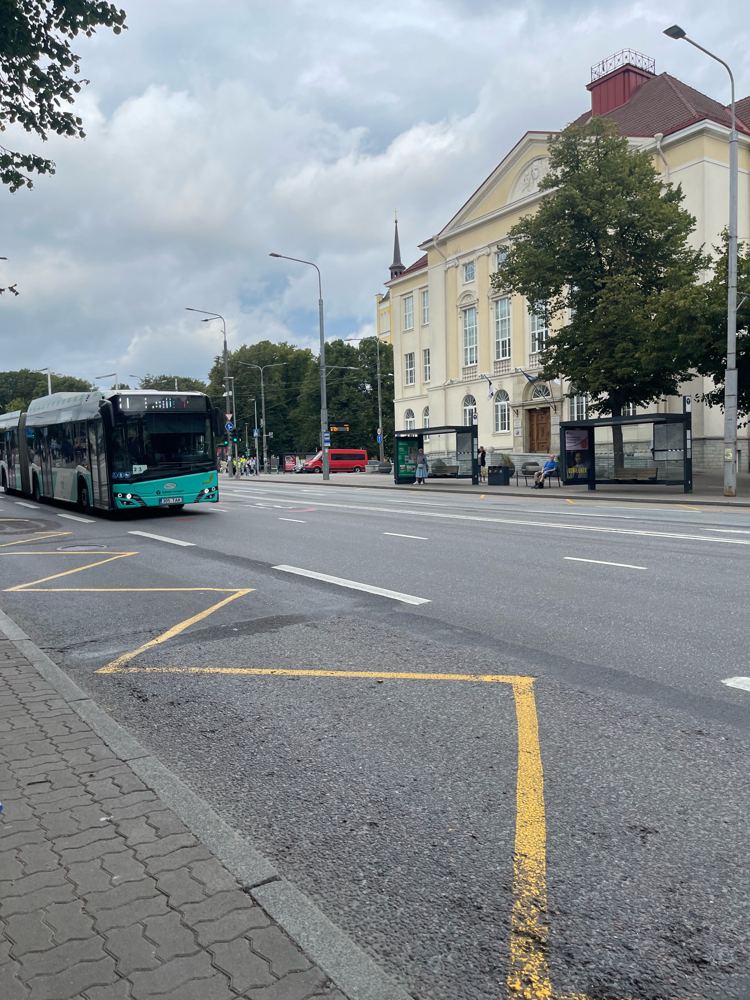 The image depicts a wide street in Tallinn, Estonia, with a modern, articulated bus in the left foreground. The bus is teal and white, displaying "23" and "305 TAK" on its front. Yellow markings are painted on the asphalt in the foreground, likely indicating a bus stop or a restricted zone.
On the right side of the street, there are two bus shelters with people waiting. One shelter has a man sitting and another has two people standing. Behind the shelters, a large, light-yellow building with a red-tiled roof and a classic facade stands. Trees line the street between the bus shelters and the building. In the background, more trees and buildings are visible under a cloudy sky. A red van is parked on the far right of the street, and a traffic light is visible. The overall scene suggests a public transportation hub in an urban setting.