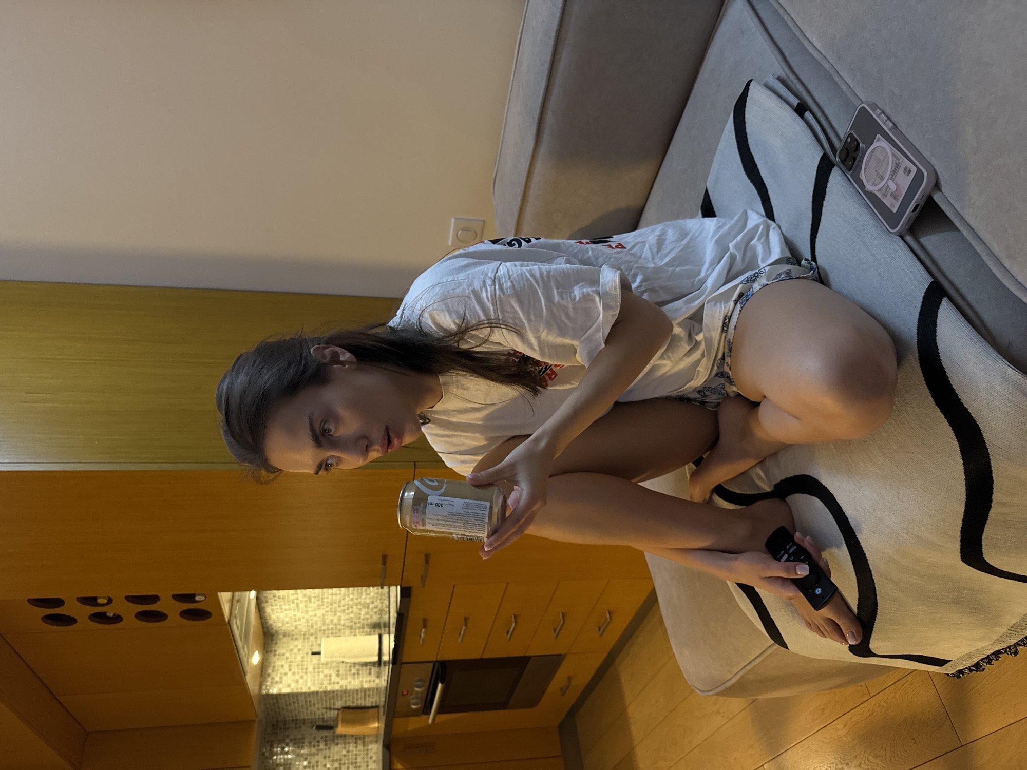 A young woman is sitting on a couch, holding a can in her right hand and a remote control in her left foot. She is wearing a white t-shirt and shorts. A phone is resting on the back of the couch. The couch has a light-colored cover and decorative pillows with black patterns. In the background, there are kitchen cabinets and appliances, including an oven and a tiled backsplash. The floor is wooden.