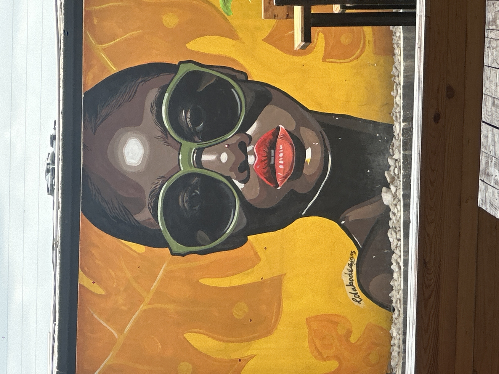 The image shows a mural of a woman wearing large, green-rimmed sunglasses and bright red lipstick. The mural is painted on a yellow and orange background with abstract leaf-like shapes. The woman's face is dark-skinned, and she has dark hair. Below the mural, there is a border of small, grey stones. In the bottom half of the image, a wooden structure is visible, possibly a bench or table, suggesting the mural is part of an outdoor seating area. The mural appears to be located in the city of Howayi, Nigeria. The overall impression is of a vibrant and artistic public space.