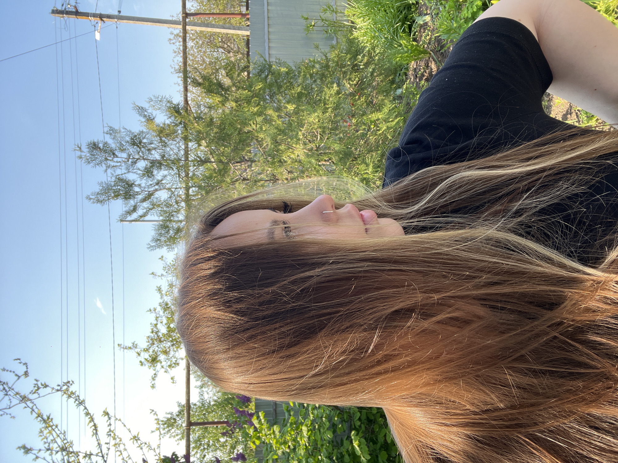 A profile view of a fair-skinned person with long, light brown hair, wearing a black top. The setting appears to be outdoors, with green foliage in the background. In the distance, utility poles and wires are visible against a blue sky. A fence is also partially visible. While the location is specified as Novopokrovskaya, Russia, there are no elements in the image to confirm it.