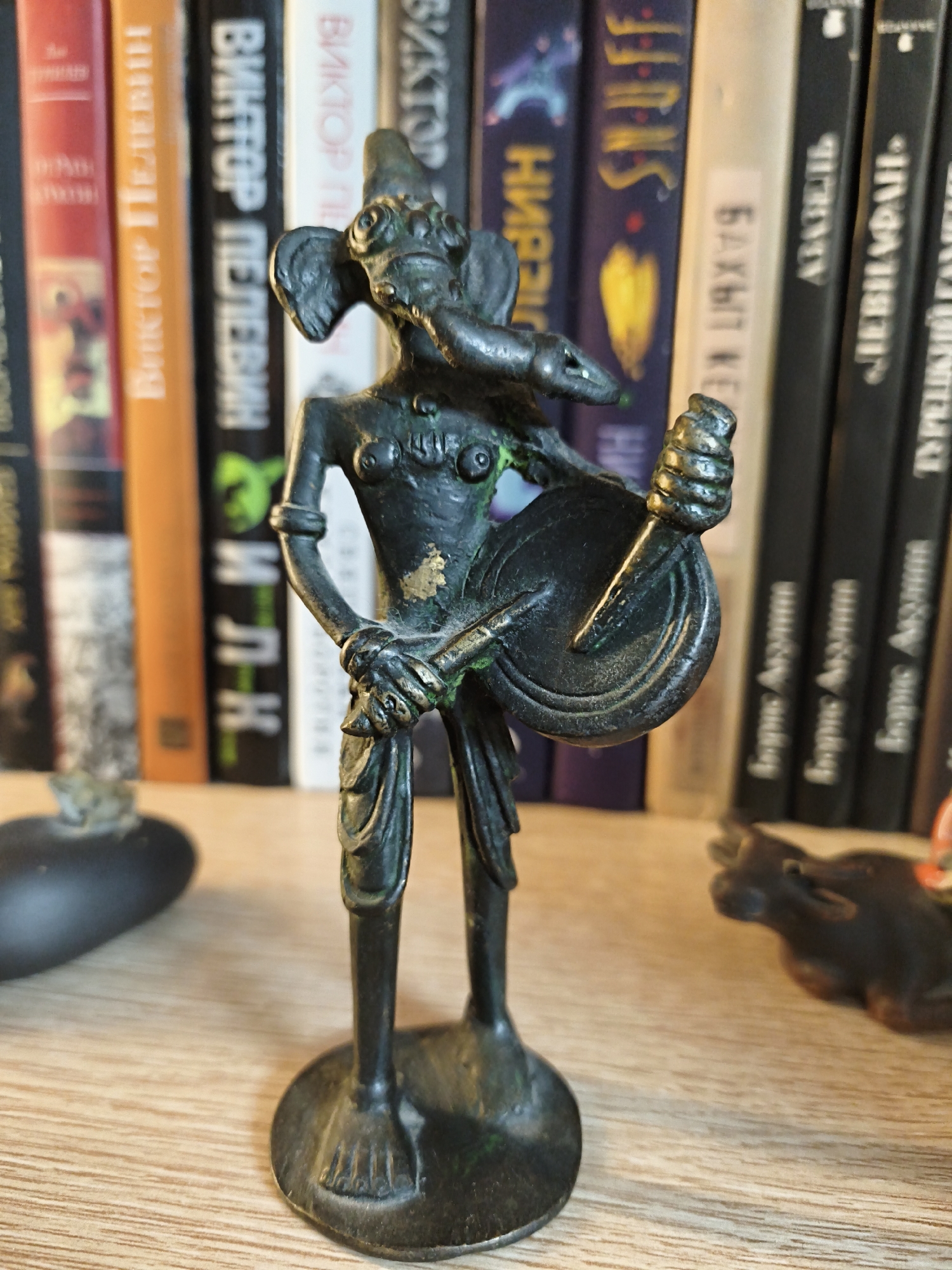 This media file is a close-up, eye-level photograph of a small, dark, bronze figurine of a deity with an elephantine head, standing on a wooden shelf. The figurine appears to be playing a stringed instrument held in its hands. It is a solitary figure, positioned centrally in the frame. Behind the figurine, a bookshelf is visible, filled with books. The spines of several books are partially legible, displaying text in Cyrillic script. Visible titles include "ВИКТОР ПЕЛЕВИН," "SNUFF," "БАКЫТ КЕР," "АЗИЗЕЛЬ," and "Борис Акулина." The lighting suggests an indoor setting, possibly a room with ambient light, casting soft shadows. The figurine exhibits signs of age or wear, with some green patina and gold discoloration in places, hinting at its material composition and possibly its age. The overall scene evokes a sense of quiet display or collection, with the figurine as the primary focus. The presence of books suggests an intellectual or cultural context, perhaps a study or library.