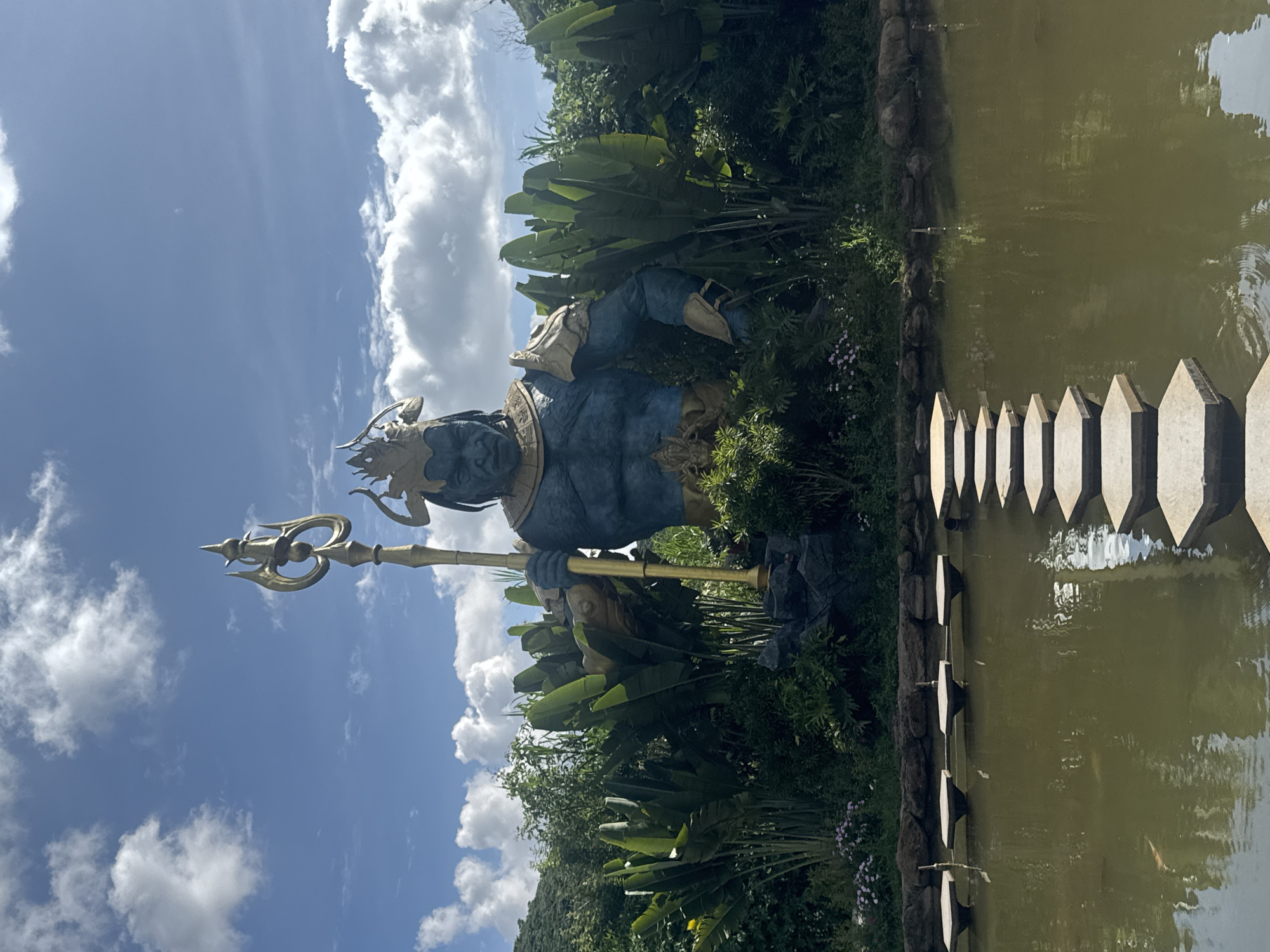 The image depicts a large, imposing blue statue of a deity holding a golden trident, situated amidst lush greenery and tropical foliage. The statue is the central focus, with its muscular build and stern expression conveying a sense of power. Behind the statue, a dense growth of large-leafed plants and trees forms a natural backdrop. In the foreground, a calm, murky pond is crossed by a pathway of hexagonal stepping stones. The sky above is a bright blue, dotted with fluffy white clouds. The scene appears to be located outdoors, possibly in a garden or temple complex, and is consistent with outdoor locations in Thôn 1, Vietnam, given the tropical vegetation. There are no people visible in the image.