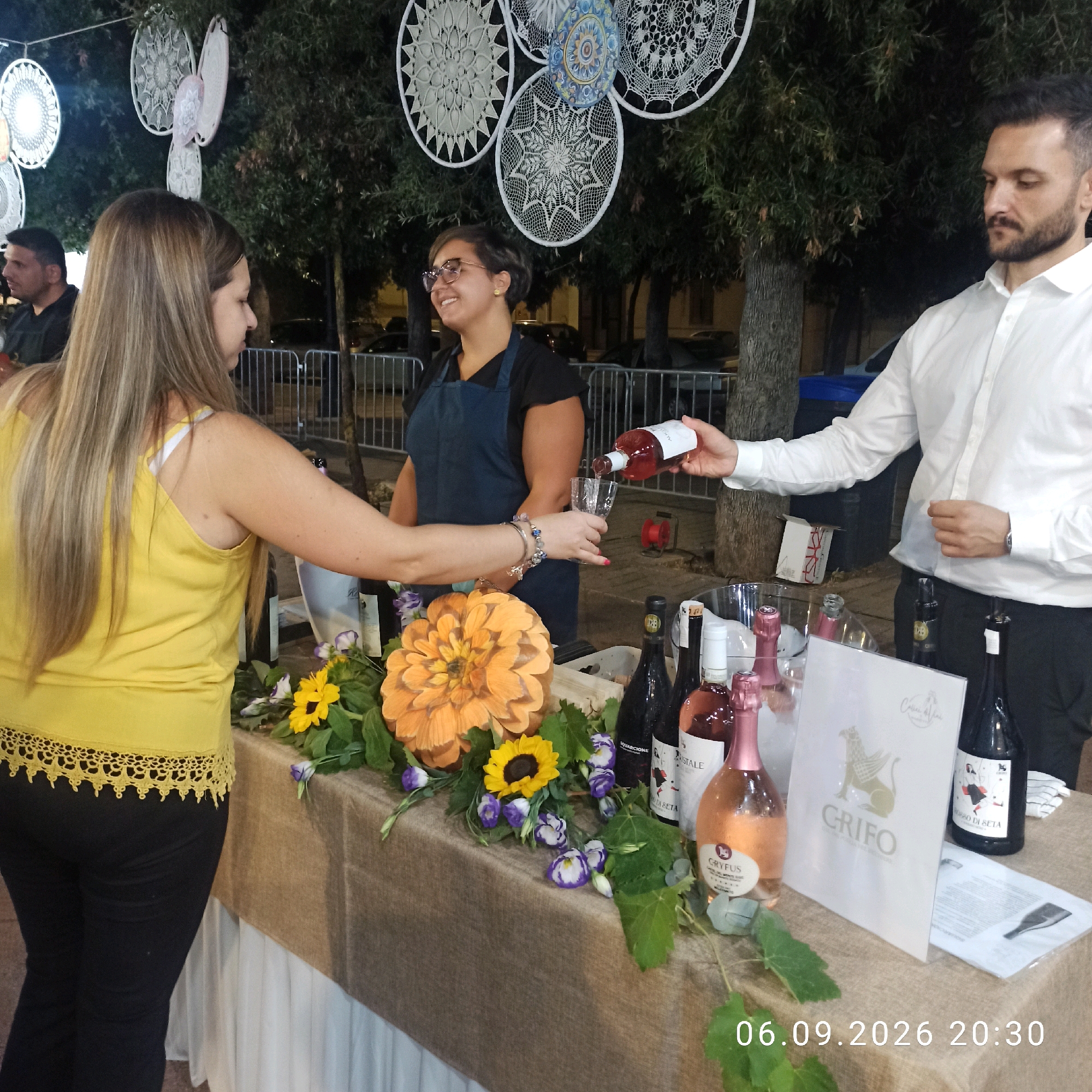 A man in a white shirt is pouring rose wine from a bottle into a glass held by a woman wearing a yellow top. They are standing behind a table laden with bottles of wine, flowers, and a decorative orange dahlia. The table is draped with a burlap cloth and a white cloth. In the background, there are trees and decorative dreamcatchers hanging from strings. The scene appears to be an outdoor event. A date stamp overlay reads "06.09.2026 20:30". The presence of a specific date suggests this might be a recorded event, though the location is not explicitly stated in the image itself, but rather inferred from external context provided by the user.