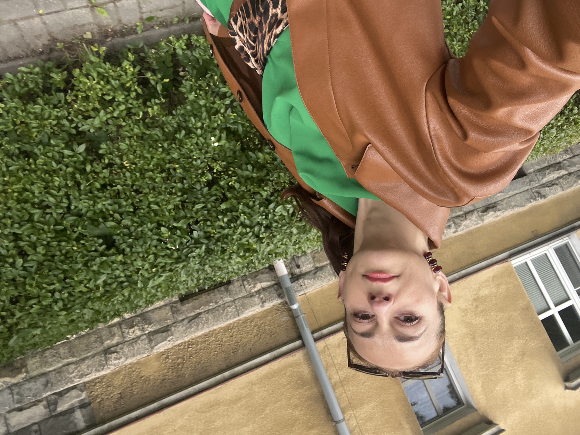 The image shows a woman outdoors, facing the camera with her head tilted. She is wearing a brown leather jacket over a green top and a leopard print fabric. She has dark hair and is wearing glasses. The background features a wall made of stone and a yellow building with windows. There is also green foliage in the background, suggesting a garden or park setting. The lighting indicates it is daytime. The woman's expression is neutral.