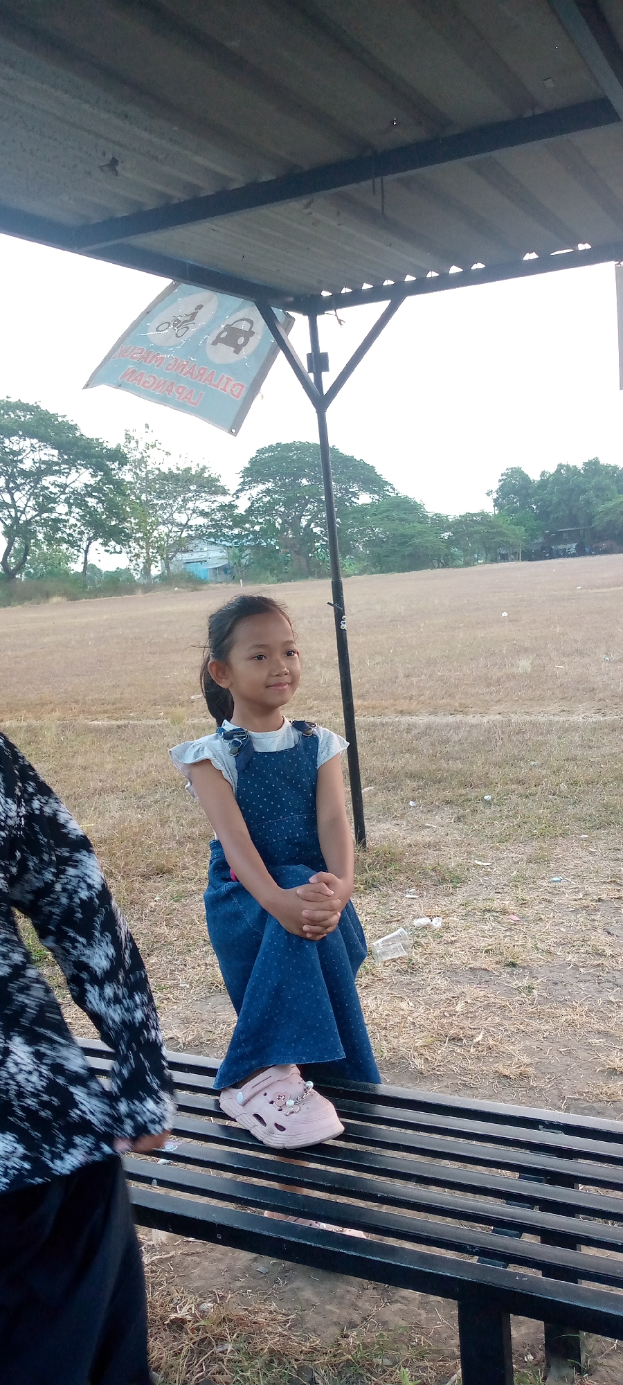 A young girl sits on a black metal bench under a covered structure, facing right and smiling. She wears a light blue shirt with short sleeves and a dark blue polka dot overall dress. Her hands are clasped in her lap, and she has light pink Croc-style shoes on her feet. To the left, a person in a black and white patterned top is partially visible, also sitting on the bench. The background shows a dry, grassy field with trees and a building in the distance. A sign is hanging from the covered structure, displaying pictograms of a bicycle and a car with a red circle and slash, indicating a prohibition. The sky is overcast. The location is the field area near the Pacing Market in Pacing, Indonesia.