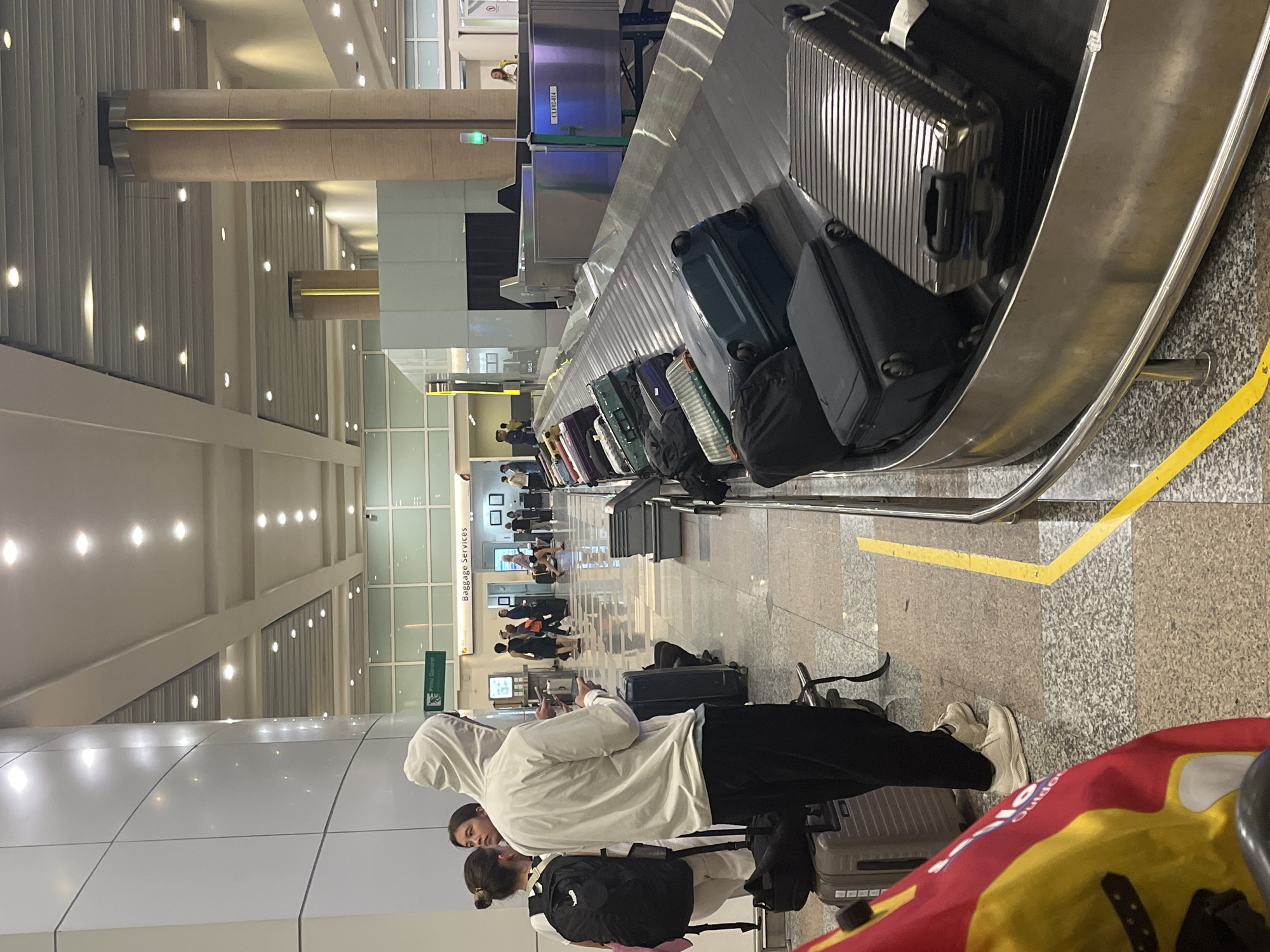 The image captures an indoor scene within a modern airport terminal, specifically a baggage claim area identified by a "Baggage Services" sign, located in Kuta, Indonesia.
In the foreground, a person wearing a white hooded jacket, black pants, and white sneakers stands next to a grey rolling suitcase, looking down at a mobile device. To their left, a person with dark hair and a black backpack is partially visible, facing away from the camera, with another individual's face partially visible between them. The floor in this area is a light-colored, mottled tile or terrazzo, marked with a distinct yellow L-shaped line on the right.
The right side of the image features a baggage carousel curving into the foreground, carrying multiple suitcases and bags. These include hard-shell cases in silver, dark blue, and green, along with several black soft bags.
In the mid-ground, behind the carousel, a baggage screening system with metallic panels and a dark intake opening is visible. A green light illuminates on a pole positioned next to this equipment, which also bears the label "CLP.02-014." Further in the background, several individuals are present, walking or standing. The terminal features tall, cylindrical pillars with vertical lighting elements. The ceiling is high, with recessed lighting and a combination of smooth panels and ribbed sections. Signs for "Pintu Darurat" (Emergency Exit) and "Baggage Services" are visible on the back wall, which also incorporates large glass windows or partitions. The environment is brightly lit and appears clean and organized.