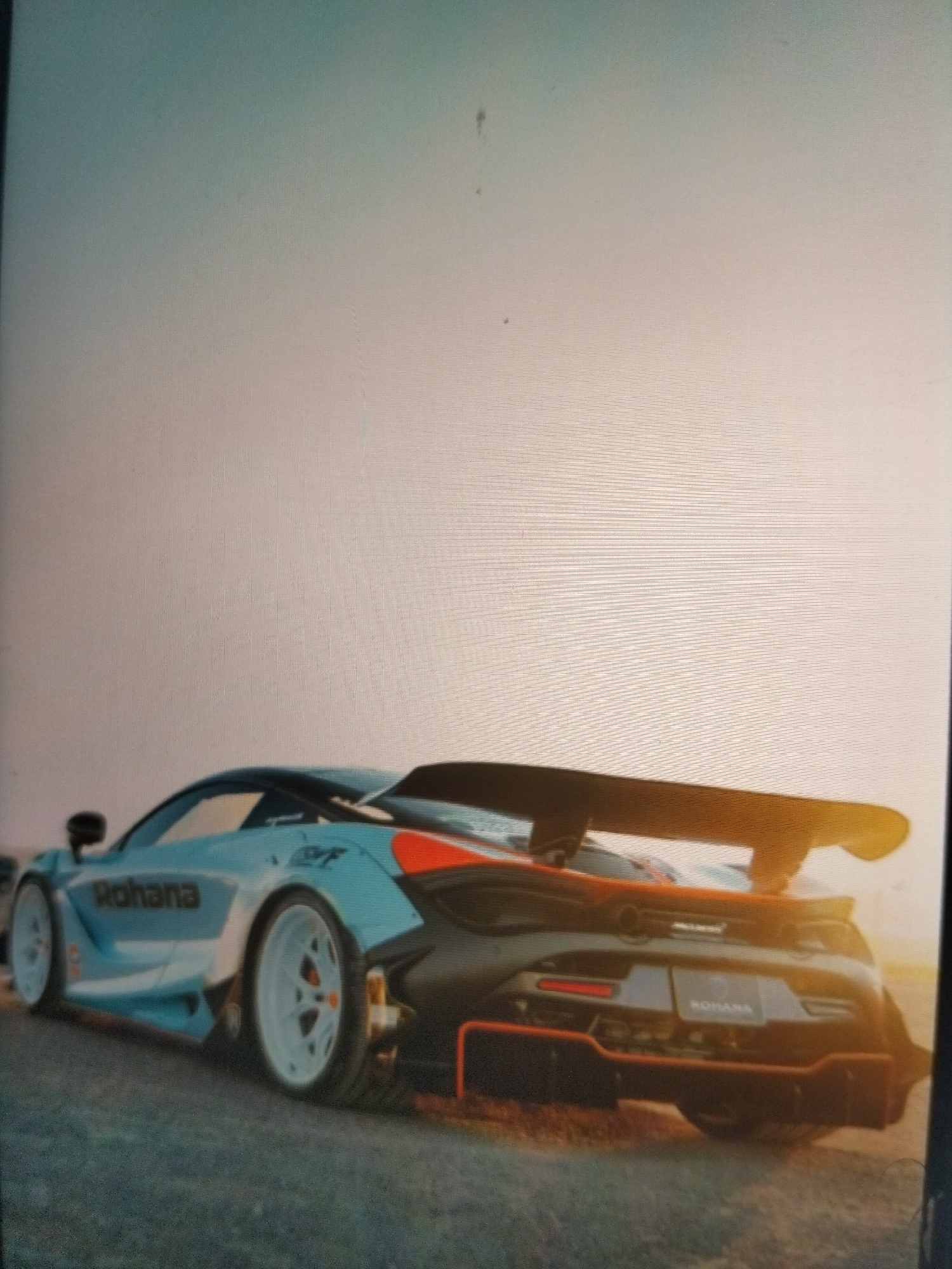 A rear, low-angle, eye-level view of a custom light blue McLaren sports car with a large black wing and red accents. The car is parked on a dark, grainy surface that could be gravel or asphalt. The sky is a gradient of pale yellow to light blue, suggesting it is either dawn or dusk. The car has "Rohana" written in black on its side, and a "Rohana" emblem is visible on the rear bumper. The McLaren logo is also visible on the rear of the car. The overall mood is one of sleek, powerful automotive design. The scene is set in Payagaboh, Indonesia.
