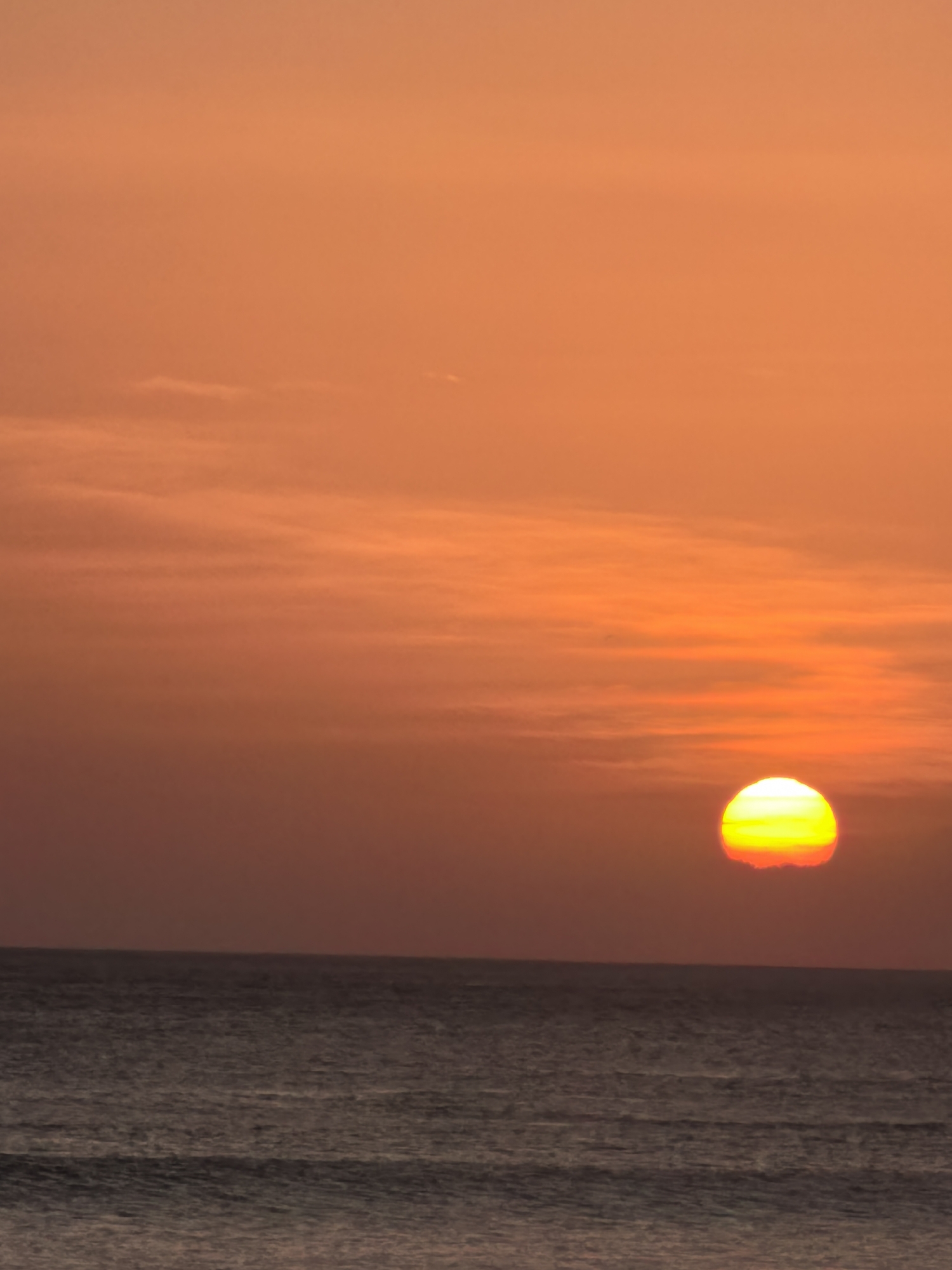 The image shows a sunset over the ocean in Kuta, Indonesia. The sun, a bright yellow-orange orb, is partially obscured by wispy clouds and is setting on the horizon. The sky is a gradient of orange and pink hues, transitioning to a darker reddish-orange at the top. The ocean below is a deep, muted blue with subtle ripples on its surface. The line where the sky meets the sea is sharp and distinct. There are no people or man-made objects visible in the frame, presenting a natural and serene seascape.
