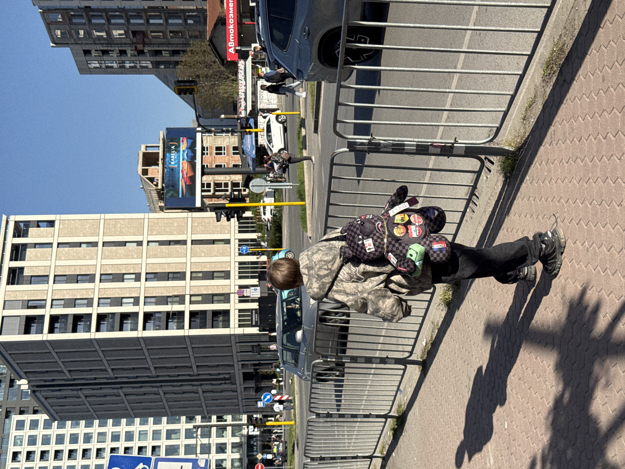 This media file captures a bright, sunny urban scene in Sofia, Bulgaria, characterized by strong sunlight and clear skies, suggesting midday or early afternoon. The image is taken from a slightly elevated perspective, looking down onto a street and sidewalk.
In the foreground, a person with short, light brown hair is prominently featured, leaning against a metal barrier that separates the sidewalk from the street. They are wearing a camouflage-patterned jacket, black pants, and dark athletic shoes. A distinctive backpack, covered in a black and white checkered pattern and adorned with numerous colorful patches and pins (including a green square, a black star, and various cartoon motifs), is worn on their back. Long, dark shadows of the person and the barrier are cast across the textured, reddish-brown paved sidewalk.
Beyond the barrier, a street with several cars is visible. A dark gray SUV is parked nearby, and a white car is further down the road. Several other people can be seen in the mid-ground, walking or standing on the sidewalk and street, appearing as indistinct figures due to the distance. Street signs, including a blue circular sign with a bicycle symbol and a red circular sign, are also present.
The background features various multi-story buildings, ranging from modern structures with horizontal window arrays to older brick buildings. Two large billboards stand out: one is blue and displays "KARELA" alongside images of people, and another red billboard clearly shows the Cyrillic text "Автокозметика" (Avtokozmetika), meaning "Car Cosmetics." Partial text "GOLAN-WARM" is also visible on a sign further back. The scene depicts a typical, bustling city environment on a clear day.