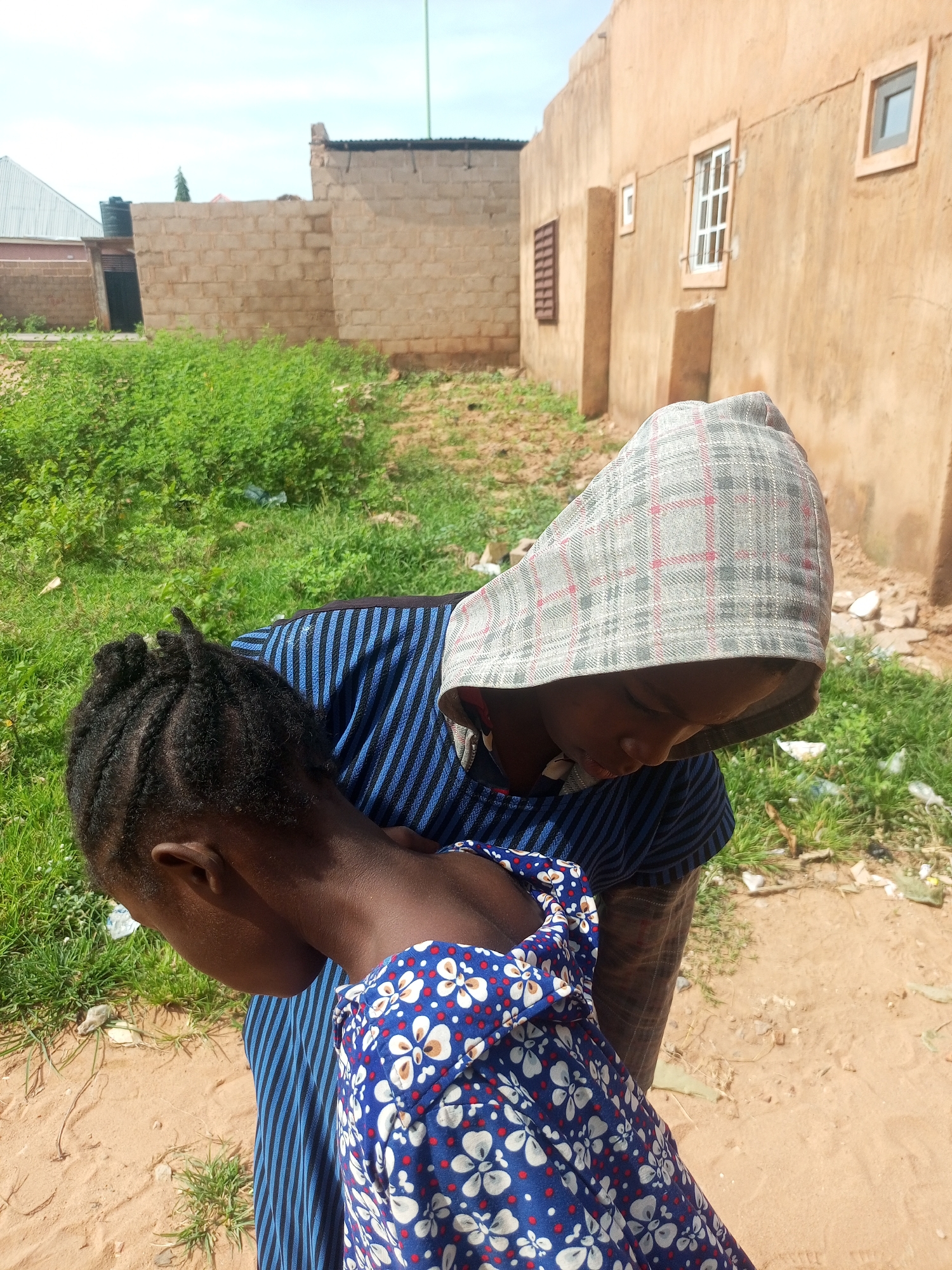 In Tempure, Nigeria, a person wearing a plaid head covering and a blue and black striped shirt leans forward, their head bowed. They are standing over another person, whose back is to the camera, and this second person is wearing a blue dress with a white floral pattern. The background consists of buildings constructed from light-colored bricks and stucco, with visible windows and doorways. Green vegetation is present in patches around the structures. The ground is covered in sand and dirt.