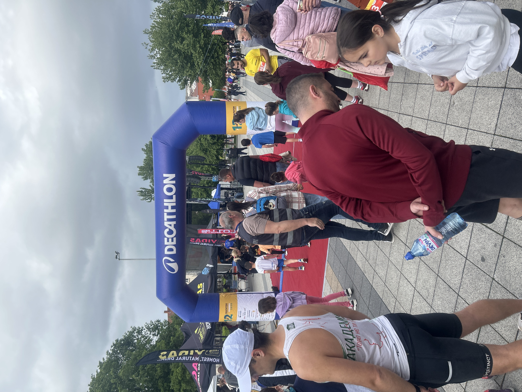 A medium-shot, outdoor photograph depicts a scene at a Decathlon marathon event in Pleven, Bulgaria, in 2026. A large, blue Decathlon archway stands near a red carpet. The sky is overcast, suggesting possible rain. A diverse crowd of people, of varying ages, is present. They are standing, walking, and interacting. One man in the foreground is wearing athletic attire, including a white cap and a tank top with "АКАДЕМИЯ" written in Cyrillic. He is looking downward. A partially visible water bottle from the brand "Devin" is held by an individual wearing a burgundy shirt and black shorts. Banners and other event paraphernalia are visible in the background, adding to the event's atmosphere.
