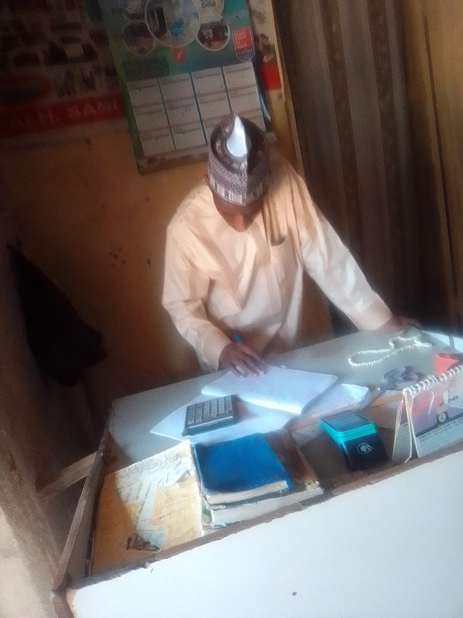 In Pantame, Nigeria, a man wearing a traditional light-colored robe and a patterned cap is seated at a desk. He is holding a pen and appears to be writing or reviewing documents on a white sheet of paper. To his right, a calculator, a stack of books, a blue notebook, a small green electronic device, a red object, and a beaded necklace are visible on the desk. A calendar with text and images is propped up on the right side of the desk, and a dark curtain hangs in the background. The environment appears to be an indoor office or workspace.