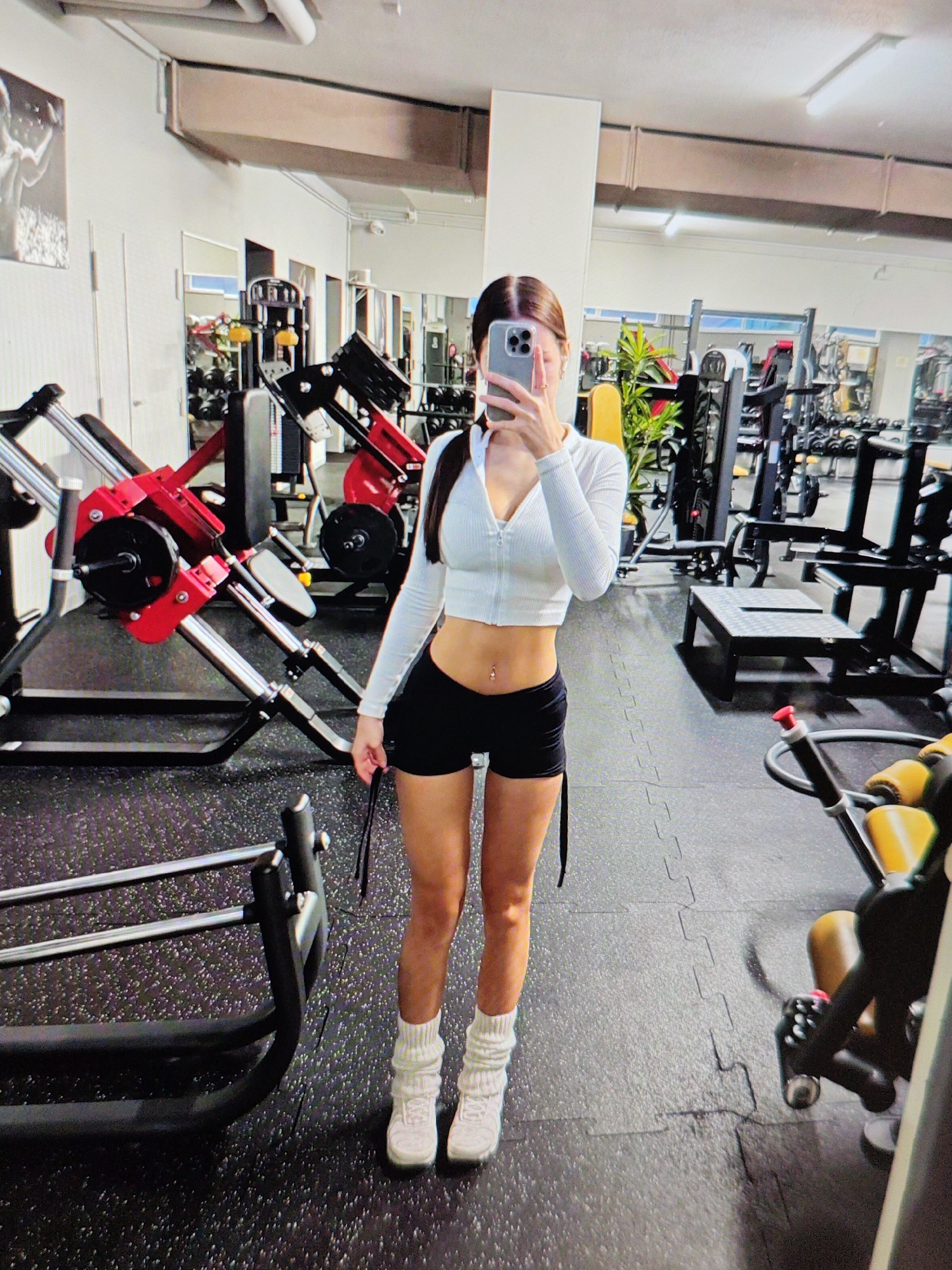 A young woman with dark hair pulled into a ponytail, wearing a white zip-up crop top, black shorts, white slouch socks, and white sneakers, stands in a gym, holding a smartphone in her right hand and taking a mirror selfie. She has a belly button piercing. The gym is equipped with various weight machines and exercise equipment, including a leg press machine with red accents. The floor is covered with black interlocking rubber mats. The environment appears to be a commercial fitness facility.