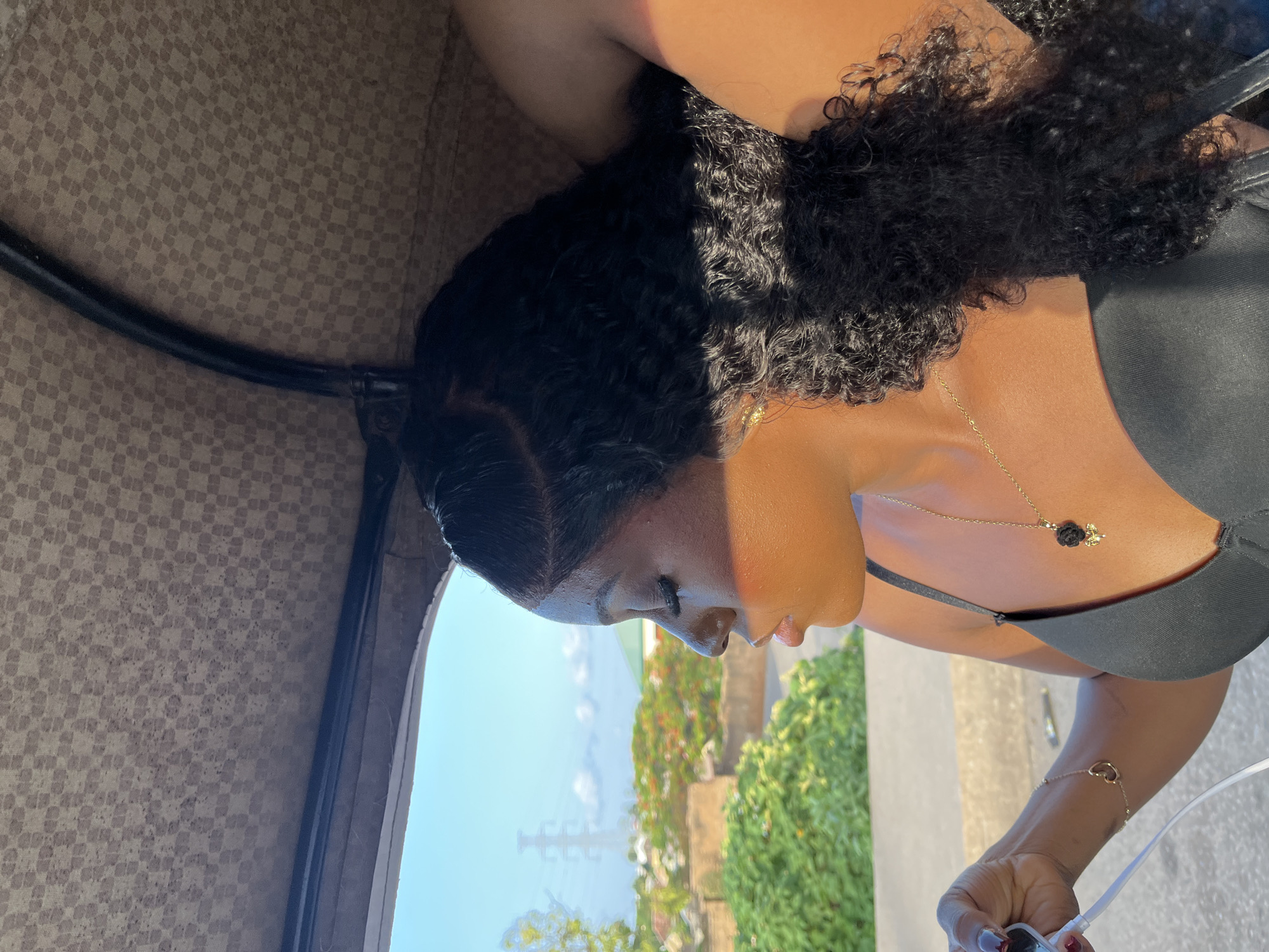 The image captures a woman in profile, facing left, seated as a passenger in a vehicle in Gura Top, Nigeria. She has dark, curly hair with visible edges, and her eyes are closed or heavily downcast. Her skin, illuminated by strong sunlight from the left, shows a warm tone. She wears dark false eyelashes and has slightly glossy lips. Adornments include a gold chain necklace with a black rose pendant and a small bee charm, a small gold hoop earring on her visible ear, and a gold heart-shaped bracelet on her left wrist. She is wearing a black top, possibly a bralette or camisole, with a strap visible. Her left hand, with dark red painted fingernails, holds a white audio cable, suggesting she is connected to a device not visible in the frame.
The vehicle's interior ceiling is a light brown fabric with a repeating dark brown checkerboard pattern, exhibiting scattered dark spots, indicative of wear or dirt. A dark, straight structural bar runs horizontally across the top of the window frame, and a curved black bar is visible on the right side of the ceiling.
Outside the vehicle, a bright, clear blue sky dominates the upper background. The visible landscape includes a variety of green foliage and trees, some displaying reddish-orange blossoms. Interspersed among the greenery are light-colored, low-rise buildings or walls. In the far distance, a tall, grey metal power transmission tower is distinctly visible against the sky. A paved road or pathway is visible immediately outside the vehicle's window in the lower-left portion of the frame.