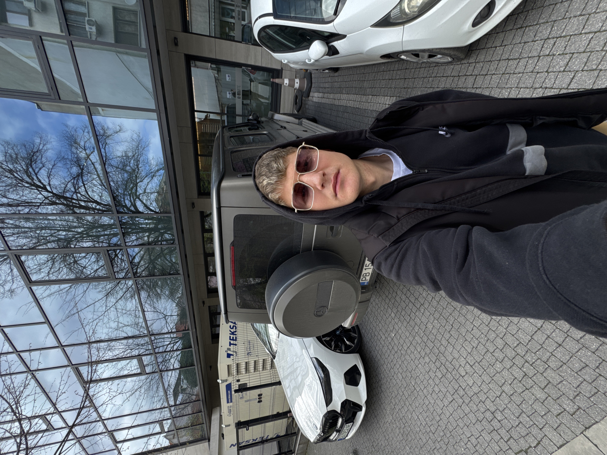 The image is a selfie taken outdoors. The person in the foreground is wearing sunglasses and a hooded jacket. Behind the person, there is a dark gray SUV with a spare tire mounted on the back. Adjacent to the SUV is a white car. A modern building with a glass facade is visible on the left, reflecting the sky and bare trees. The ground is paved with gray cobblestones. A sign with "TEKSA" is visible, suggesting the location. Given the license plate "PB 15" on the SUV, the presence of "TEKSA," and the architecture in the reflection, the location is likely near the TEKSA petrol station at бул. „Санкт Петербург“ 158, 4003 Plovdiv, Bulgaria.