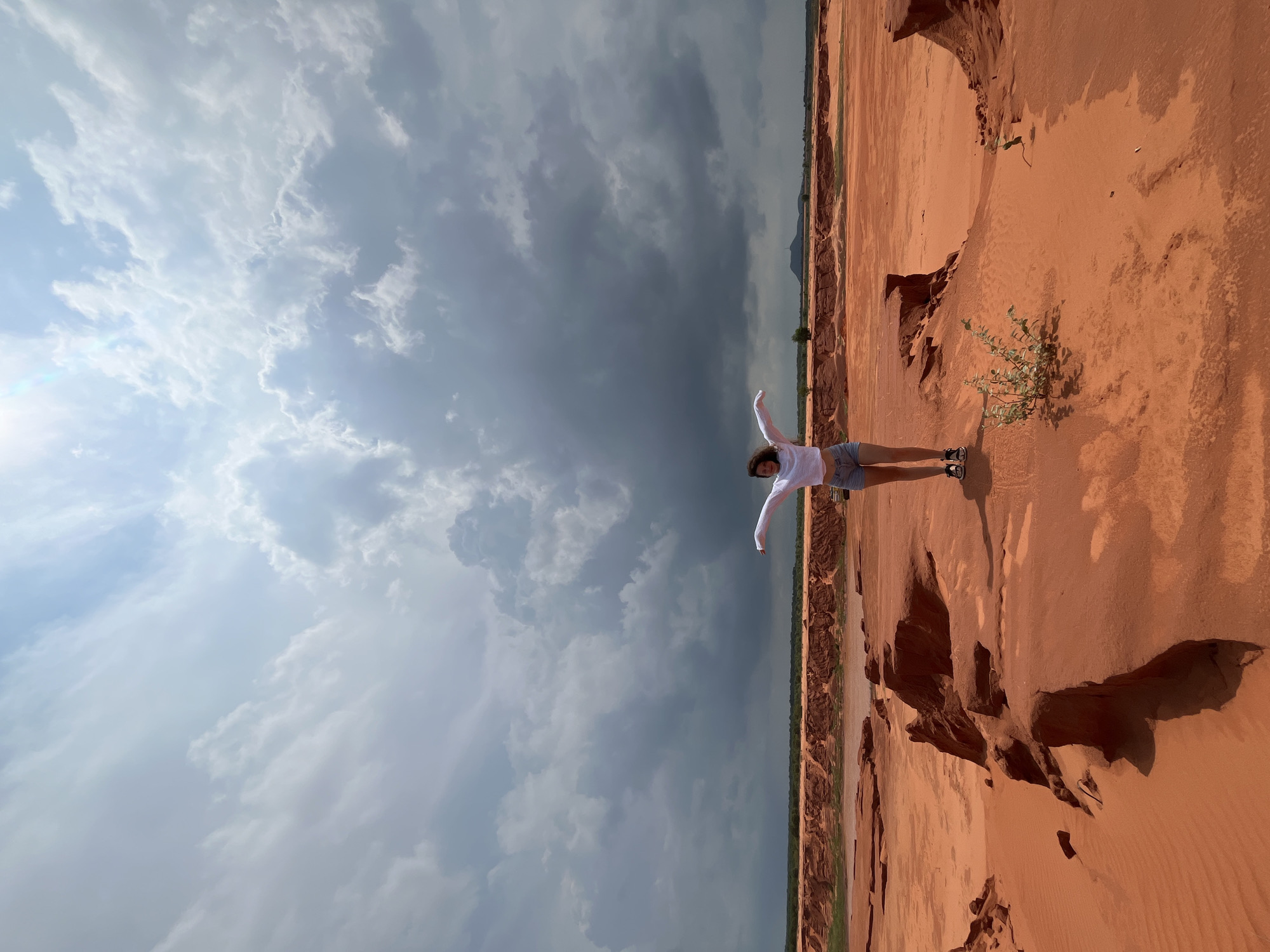 A lone young woman with dark hair stands in the center of a vast, arid landscape. She is wearing a white long-sleeved shirt and denim shorts, with her arms outstretched to the sides. The ground is covered in red sand, with some eroded rock formations. The sky above is dramatic, with heavy, dark clouds gathering on the horizon, contrasting with a brighter, sunlit area in the upper right corner. The landscape is characteristic of the red sand dunes of Mui Ne, Vietnam.