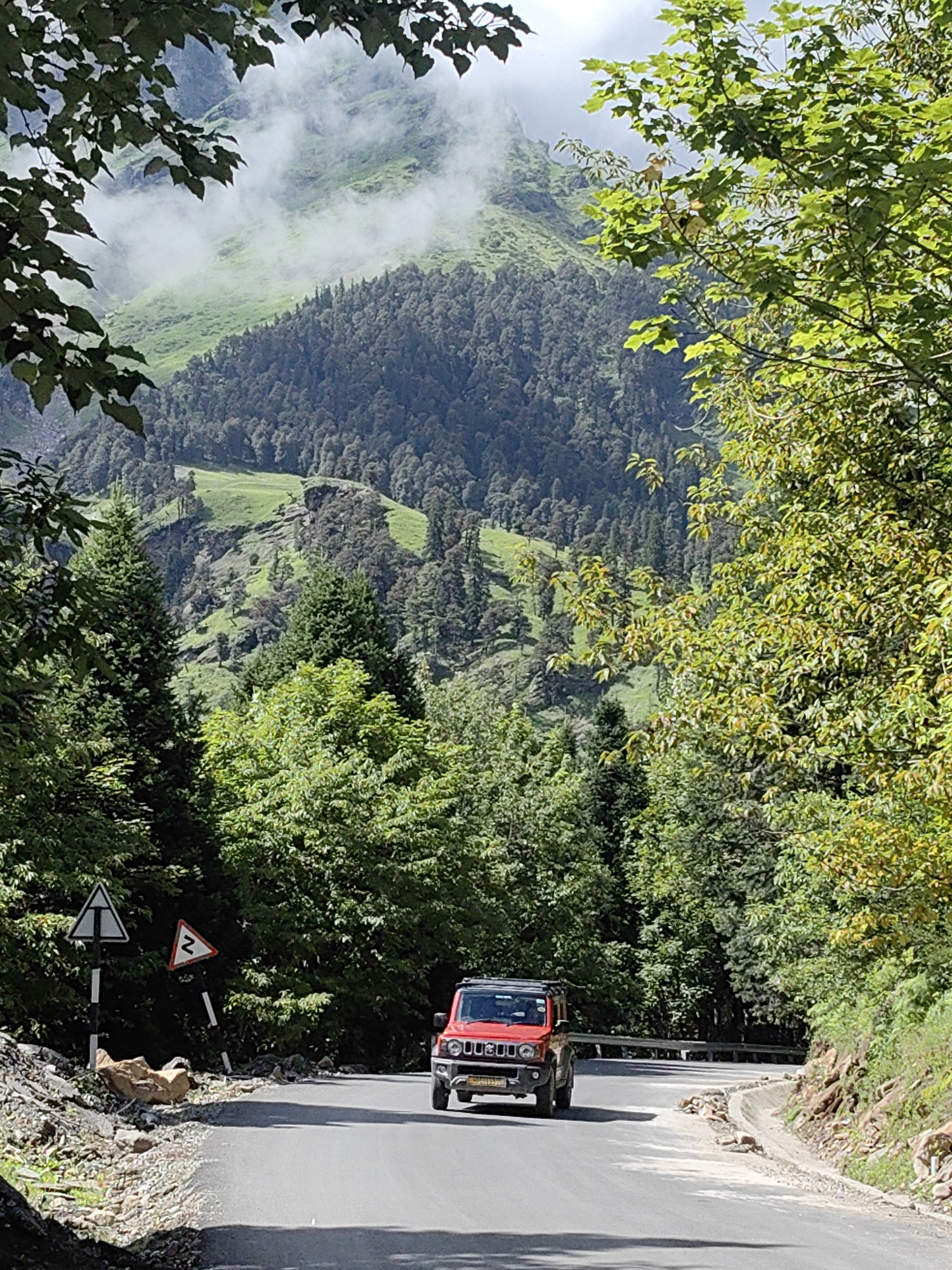 A bright red, small SUV is driving on a paved mountain road in Kothi, India. The road is winding and bordered by lush green trees on either side. In the background, steep, tree-covered mountains rise, with patches of mist clinging to the slopes. The sky is partly cloudy. Two traffic signs are visible on the left side of the road, one triangular and white with a black border and symbol, and another triangular and white with a black border and a black zig-zag symbol. Rocks and debris are scattered along the roadside.