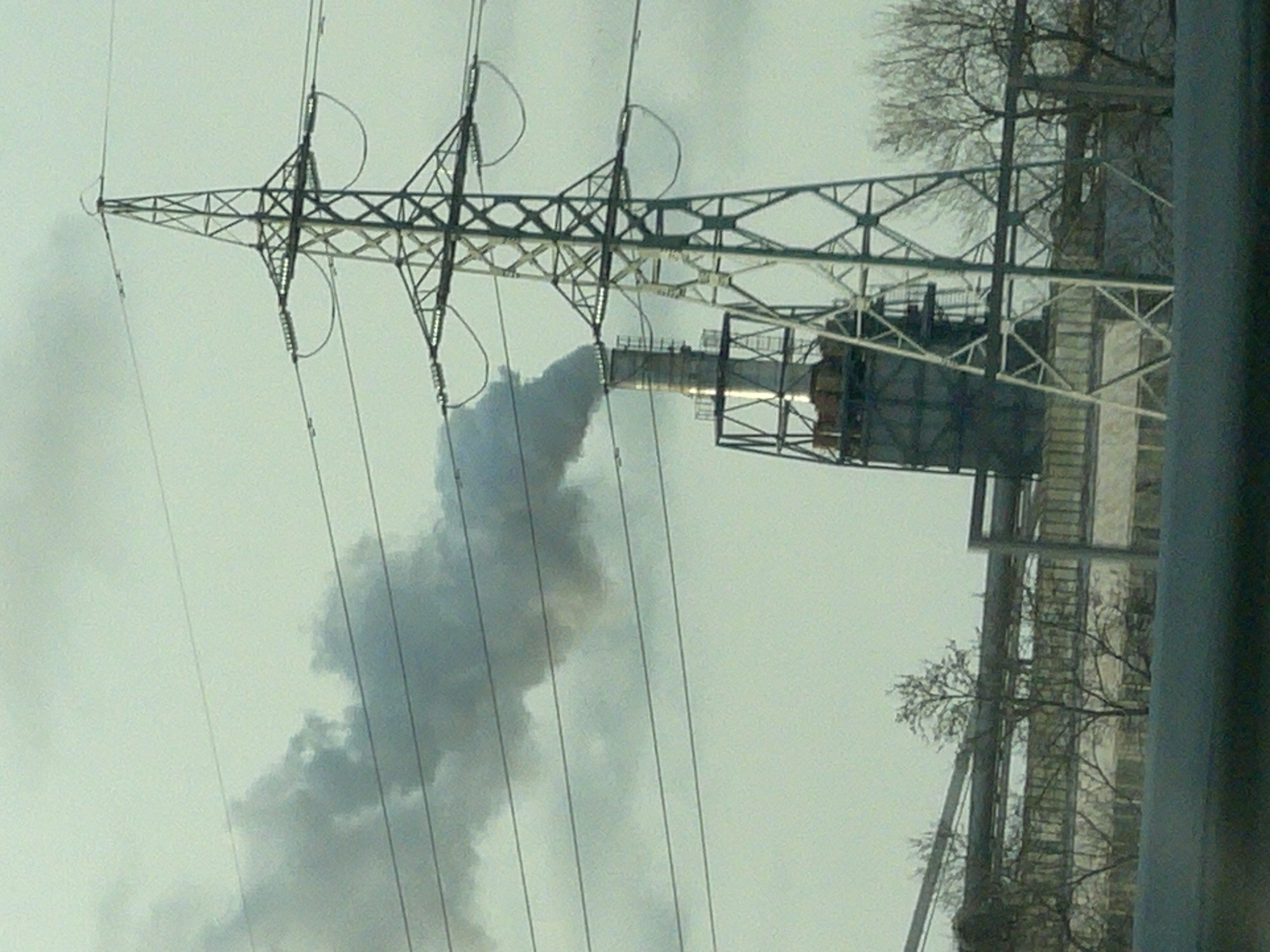 The image shows a smokestack emitting smoke in Burgas, Bulgaria. The smokestack is attached to a structure with ladders and metal framing. Power lines and a power transmission tower are in the foreground. Bare trees are visible behind the structure. The sky is overcast.