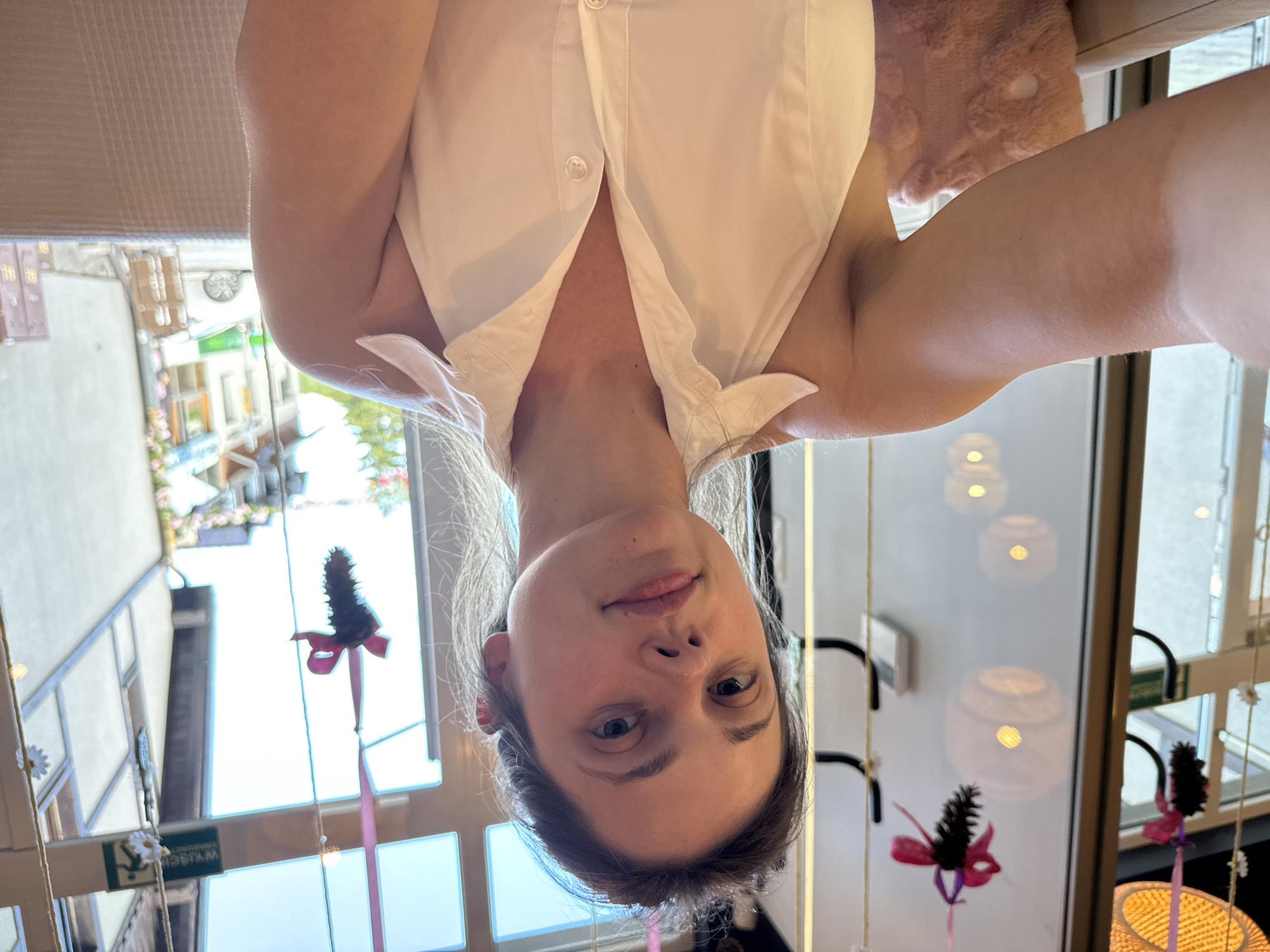 In Szklarska Poręba, Poland, a woman with brown hair pulled back sits in what appears to be a cafe or a similar establishment with large windows. She is taking a selfie, her face angled towards the camera. She is wearing a white halter top with a collar. In the background, the windows offer a view of a street scene with buildings and greenery. Decorative pinecones with pink ribbons hang from strings near the window. The interior of the establishment is partially visible, with what looks like a plush pink cushion on a chair in the foreground.