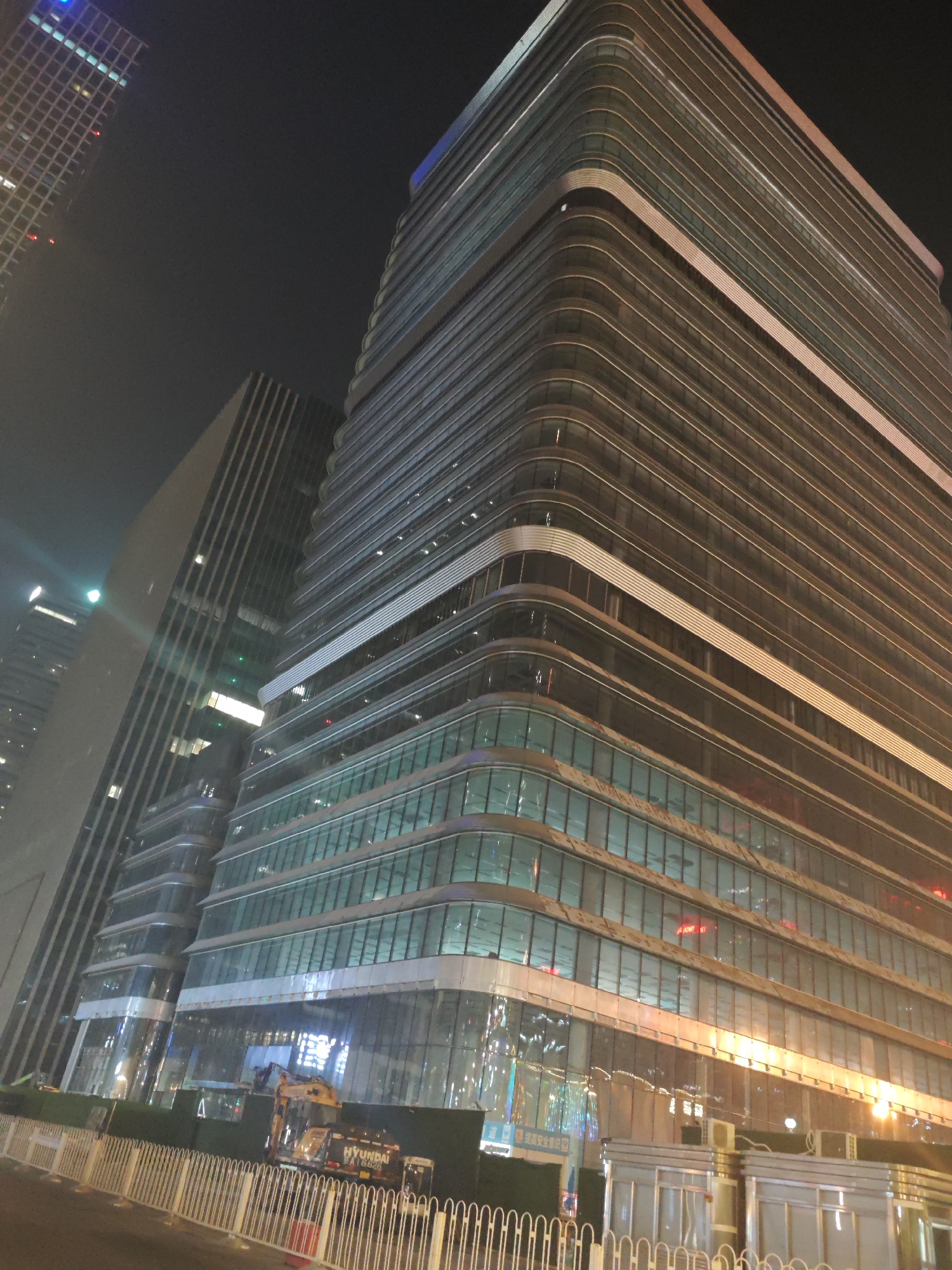 This media file captures an urban night scene in what is identified as Chaoyang District, China, featuring modern skyscrapers and a ground-level construction or renovation site.
The most prominent subject is a tall, contemporary skyscraper on the right, characterized by its curved corners and a facade composed of alternating horizontal bands of reflective glass panels and light-colored opaque sections. Many windows display internal lighting, casting yellow, white, and occasional red glows. To the left, a second, darker high-rise building with a more rectilinear design is partially visible, also with illuminated windows. Further in the upper left background, part of another, taller skyscraper with a gridded facade and blue and red accent lights can be seen.
At ground level, a white metal barrier fence delineates a work zone in front of the main building. Behind this fence, a yellow Hyundai excavator is clearly visible, parked, indicating ongoing construction or maintenance activity. The text "HYUNDAI" and "R160LC-9" is legible on the side of the excavator. Several temporary structures or utility boxes are also present near the building's base.
The time of day is clearly night, with the dark sky providing a backdrop for the brightly illuminated buildings and work area. The scene is devoid of visible people. The architecture suggests a modern, bustling urban center.