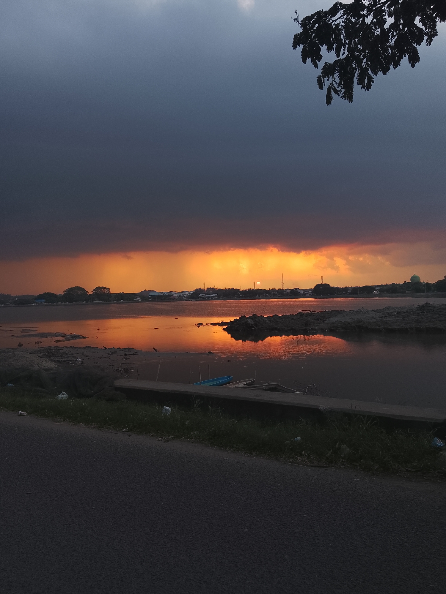 A body of water reflects a dramatic sunset with dark clouds overhead and a bright orange glow on the horizon. In the distance, a silhouetted cityscape includes buildings, trees, and what appears to be a dome-shaped structure, possibly a mosque, indicating an urban or semi-urban environment. The foreground shows a paved road and a grassy embankment with some debris, suggesting a roadside view. The scene captures a moment of natural beauty at dusk in Lhokseumawe, Indonesia, with the interplay of light and shadow creating a striking visual.