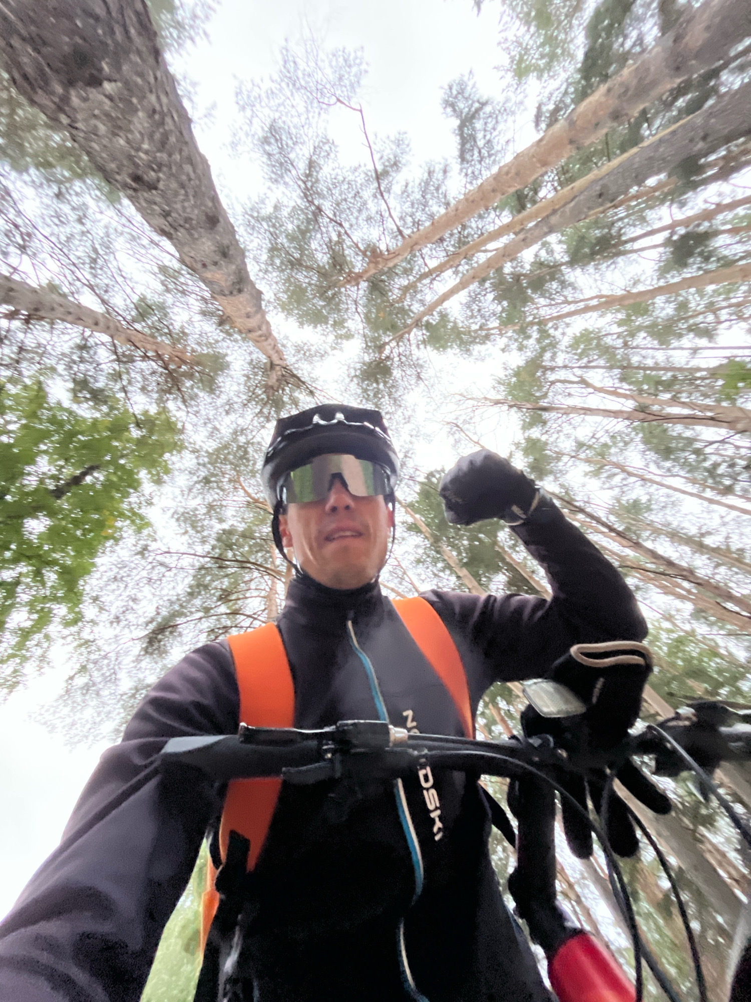A male cyclist, wearing a black helmet and oversized mirrored sunglasses, poses for a selfie while riding a mountain bike through a forest with tall, thin trees and a cloudy sky. He is wearing a black jacket with an orange vest underneath, and black gloves. He flexes his right bicep towards the camera. The handlebars of the bike are visible, with a black bag attached. The image is taken from a low angle, looking up towards the cyclist and the trees. The location is identified as Chaikovsky, Russia.