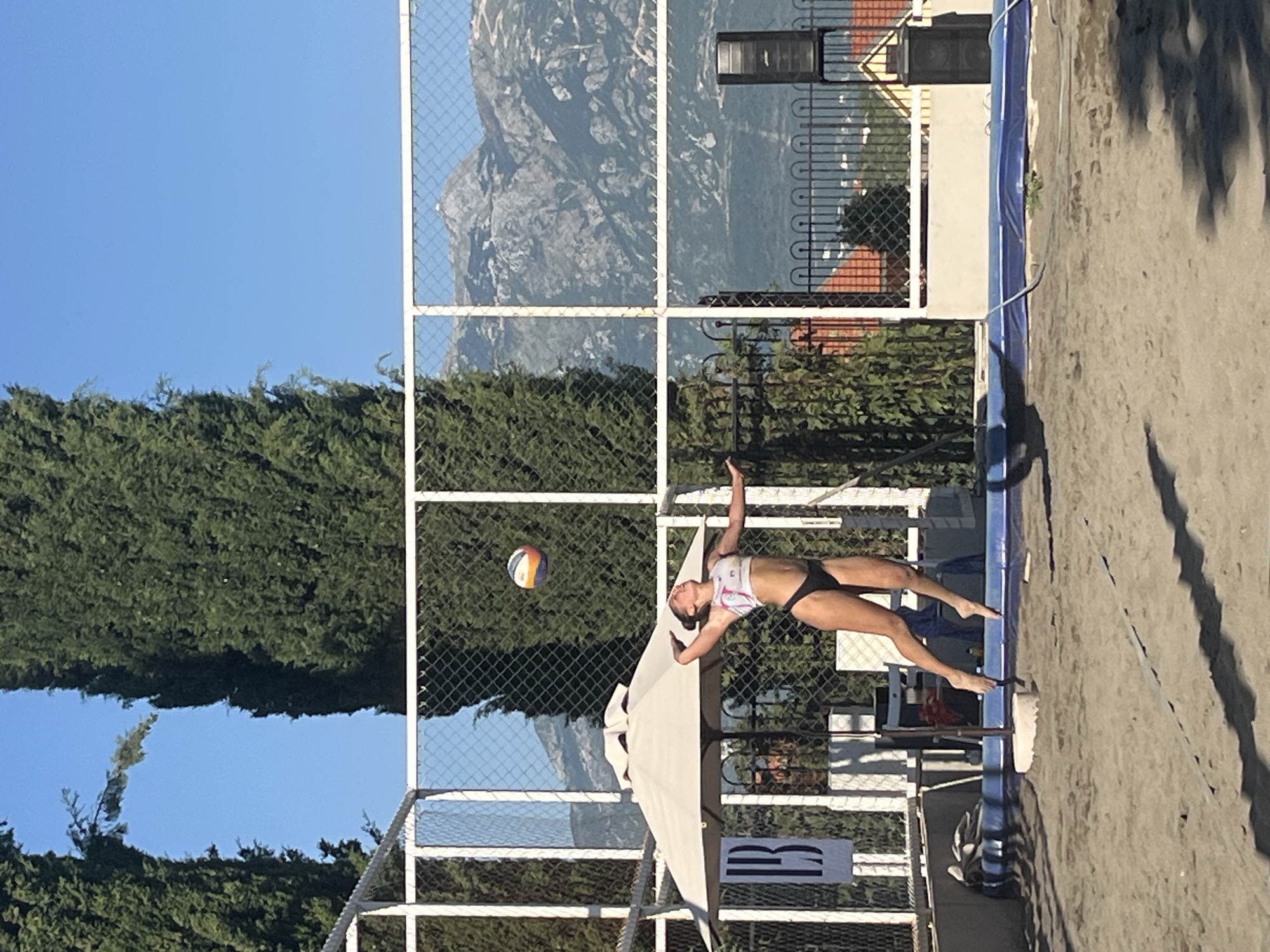 A female beach volleyball player is captured mid-air during a game on a sandy court. She is wearing a black bikini and a pink and white top. The volleyball is in the air above her head, ready to be hit. In the background, a white net encloses the court, with tall green trees and mountainous terrain visible beyond. A white umbrella provides shade to the side, and a speaker is mounted on a stand to the right. The scene appears to be set in Gošići, Montenegro, a location known for its coastal beauty and outdoor recreational facilities.