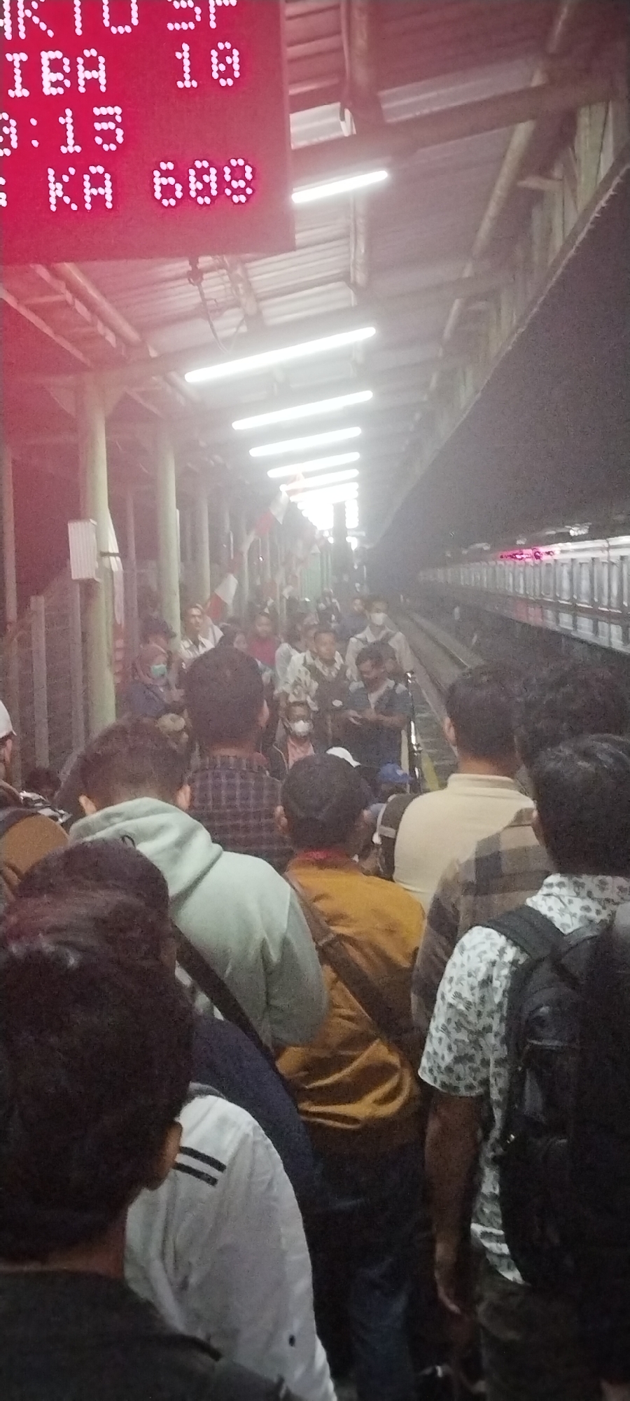 The image depicts a crowded train platform at Daru Station, Indonesia. A digital display board is prominently featured, showing train information in red LED lights. The board displays "JAKARTA SP", "CIBA 18", and "03:13 KA 609". Numerous individuals, predominantly men, are visible, many wearing face masks. They are standing and waiting, appearing to be passengers preparing to board or disembark a train, though no train is clearly visible in the frame. The environment is an indoor or covered platform with fluorescent lighting. The crowd is dense, with people standing close together. Flags are visible hanging along the platform's edge, possibly indicating a special event or national celebration. The overall atmosphere suggests a busy public transport hub.