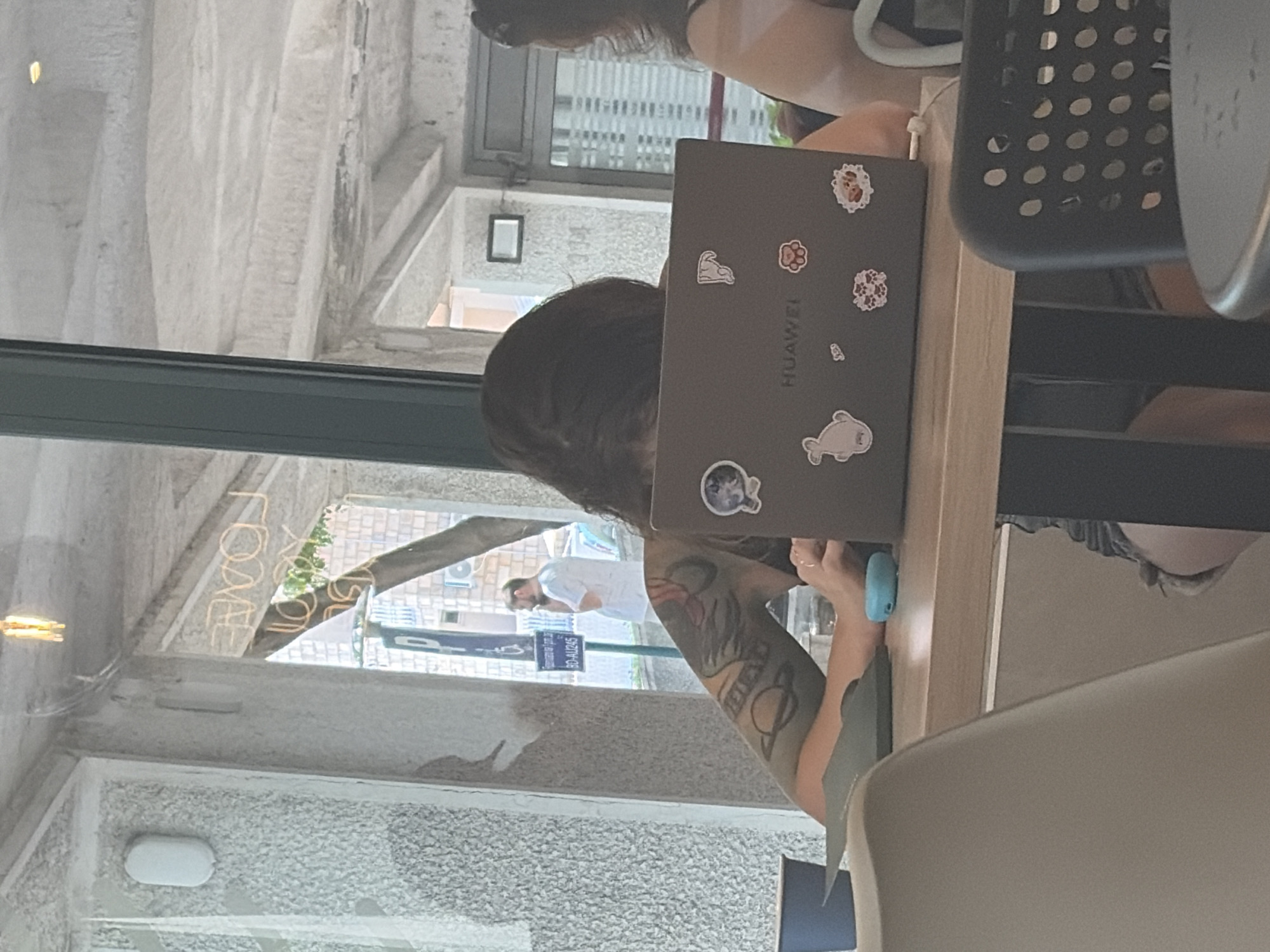 A person is working on a Huawei laptop at a cafe in Budva, Montenegro. The person has a large tattoo on their left arm, which is visible. The laptop is decorated with several stickers. In the background, a man in a white shirt is standing outside. The cafe appears to have a modern, minimalist design with concrete walls and a wooden table. The scene is viewed from behind the person working on the laptop.