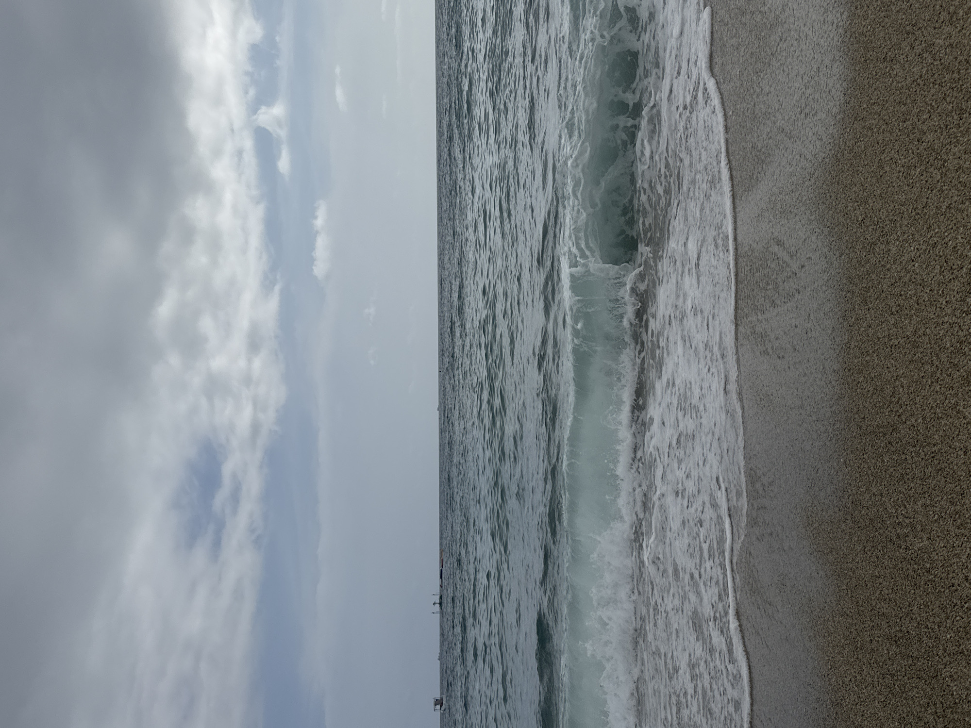 The image depicts a beach scene in Alanya, Turkey. The frame is divided vertically, with the left side showcasing a cloudy sky and the right side revealing the sea meeting the shore. Waves are breaking along the shoreline, creating white foam. The sand on the beach appears to be a dark, granular texture. In the distance, small structures or boats can be faintly discerned on the horizon.