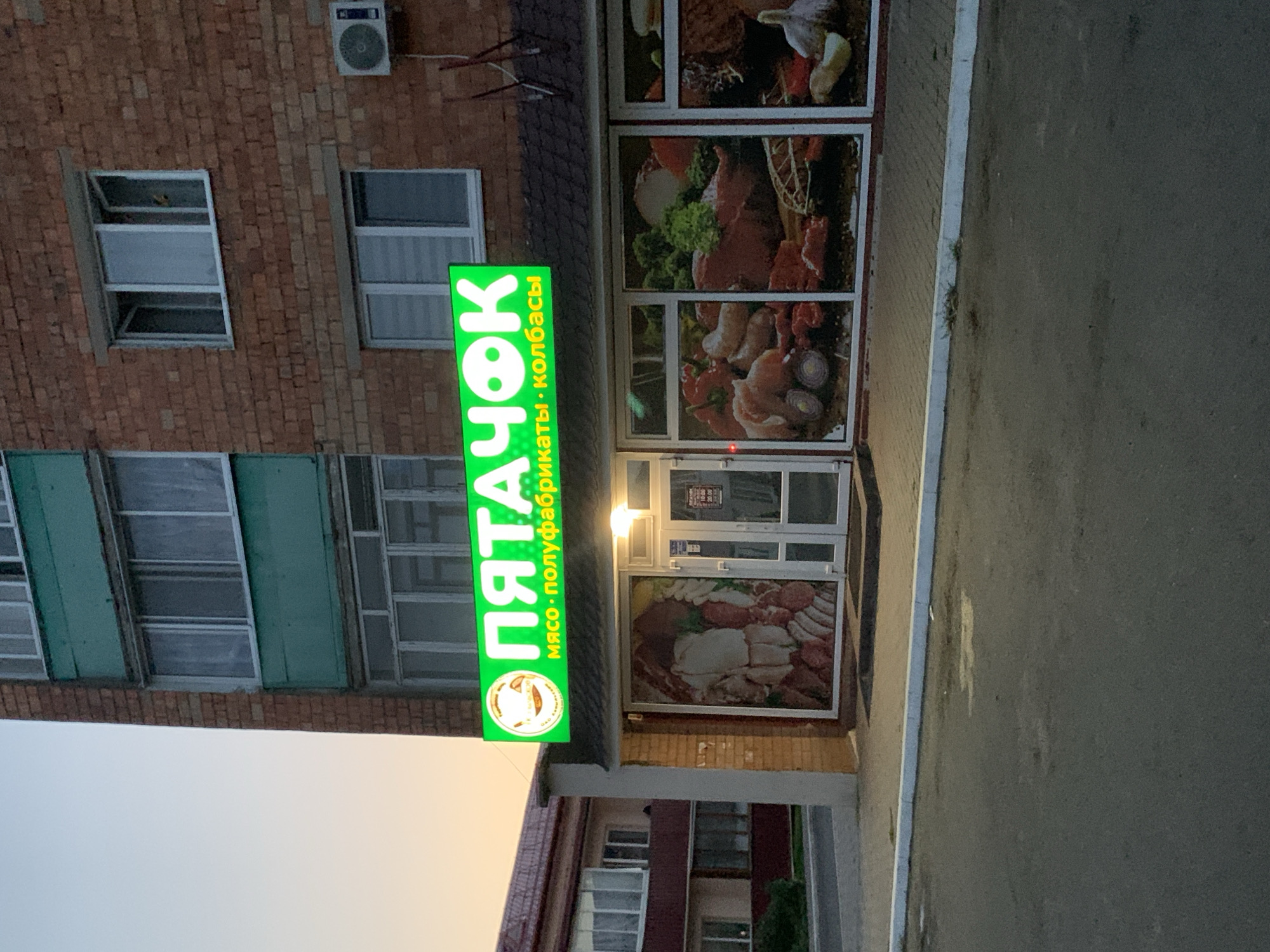 The image shows a storefront for a meat shop in Kapyĺ, Belarus. The shop's sign is bright green and reads "ПЯТАЧОК" (Pyatachok), with the smaller text below saying "мясо · полуфабрикаты · колбасы" (meat · semi-finished products · sausages). The storefront windows display images of various meats, vegetables, and sausages. The building is made of brick, with several windows visible on the upper floors. The sky is a pale orange, suggesting it is either early morning or late evening. The street in front of the shop is paved with asphalt. No people are visible in the image.