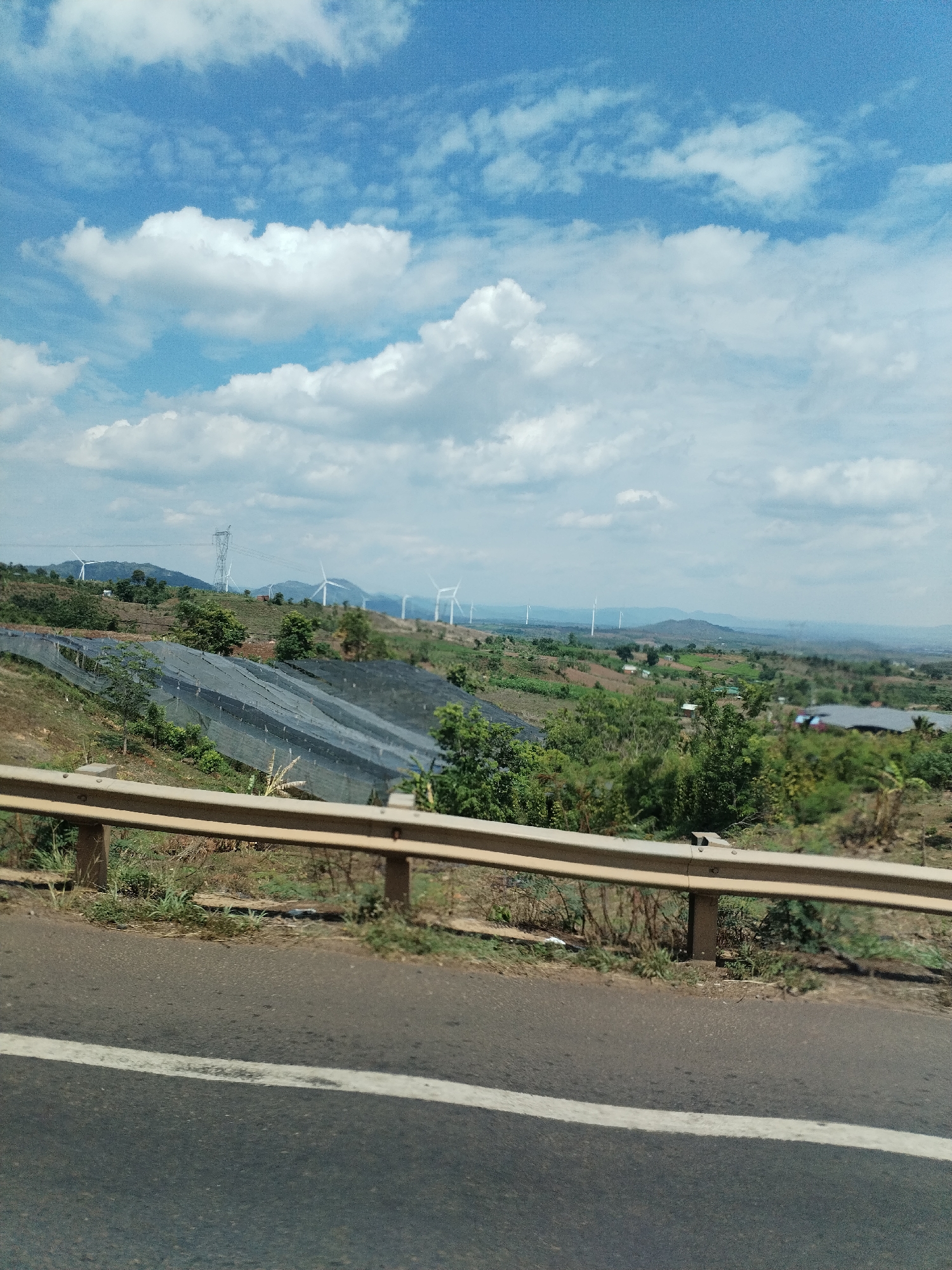 The image shows a roadside view in what appears to be a rural area of Thôn 4, Vietnam. The foreground features an asphalt road with a white lane marking and a metal guardrail. Beyond the guardrail, the terrain slopes downward, revealing a landscape of green vegetation, including trees and crops. A section of the hillside is covered with dark netting. In the distance, a series of wind turbines are visible against a backdrop of hazy mountains and a partly cloudy sky. An electrical transmission tower can also be seen on the ridge.