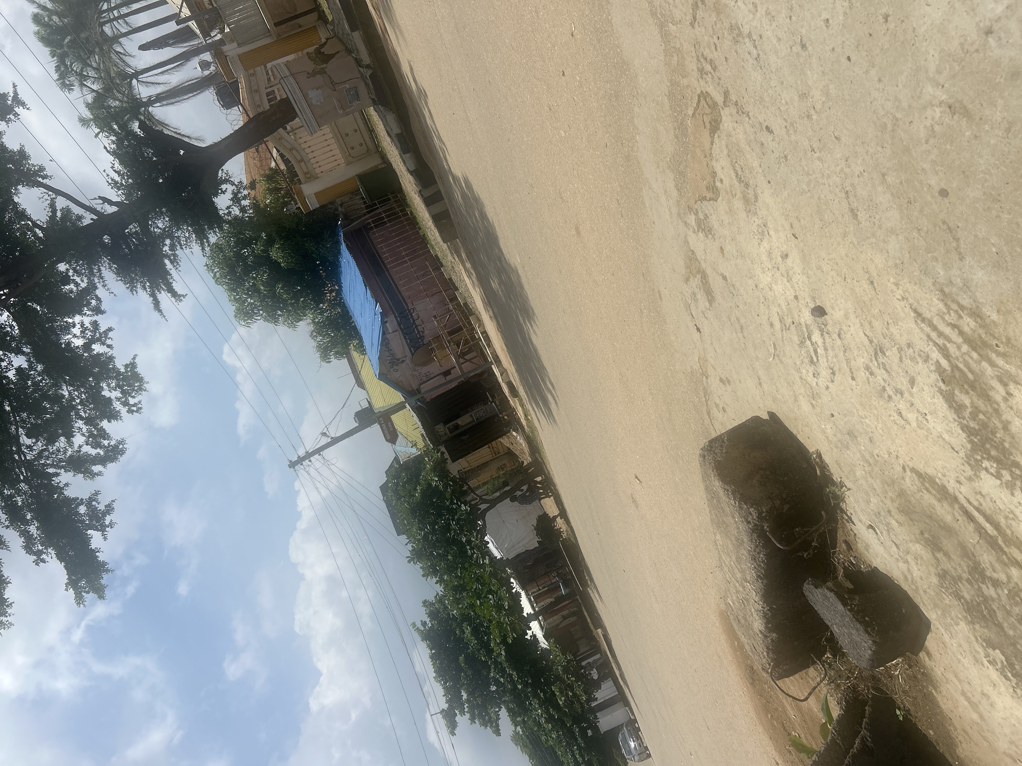 The image depicts a street scene in Abuja, Nigeria, on a cloudy day. The foreground is dominated by a dusty, light-colored road with two large, dark, rough-hewn stones in the lower left corner. The stones appear weathered and overgrown with small patches of dry grass.
Further down the road, on the right side, is a building with a yellow facade and a prominent blue corrugated metal roof. A white fence with decorative yellow posts lines the front of the property. To the left of this building, there are several other structures, some with corrugated iron roofs and others appearing to be shops or market stalls. Green trees with dense foliage are visible throughout the scene, casting shadows on the road.
In the background, power lines stretch across the sky, supported by utility poles. Palm trees are also present, suggesting a tropical or sub-tropical climate. A silver car is parked on the left side of the road in the distance. The overall impression is one of a typical urban street in a developing area, with a mix of residential and commercial buildings and a lot of vegetation. The atmosphere is generally casual, with no obvious signs of distress or emergency.