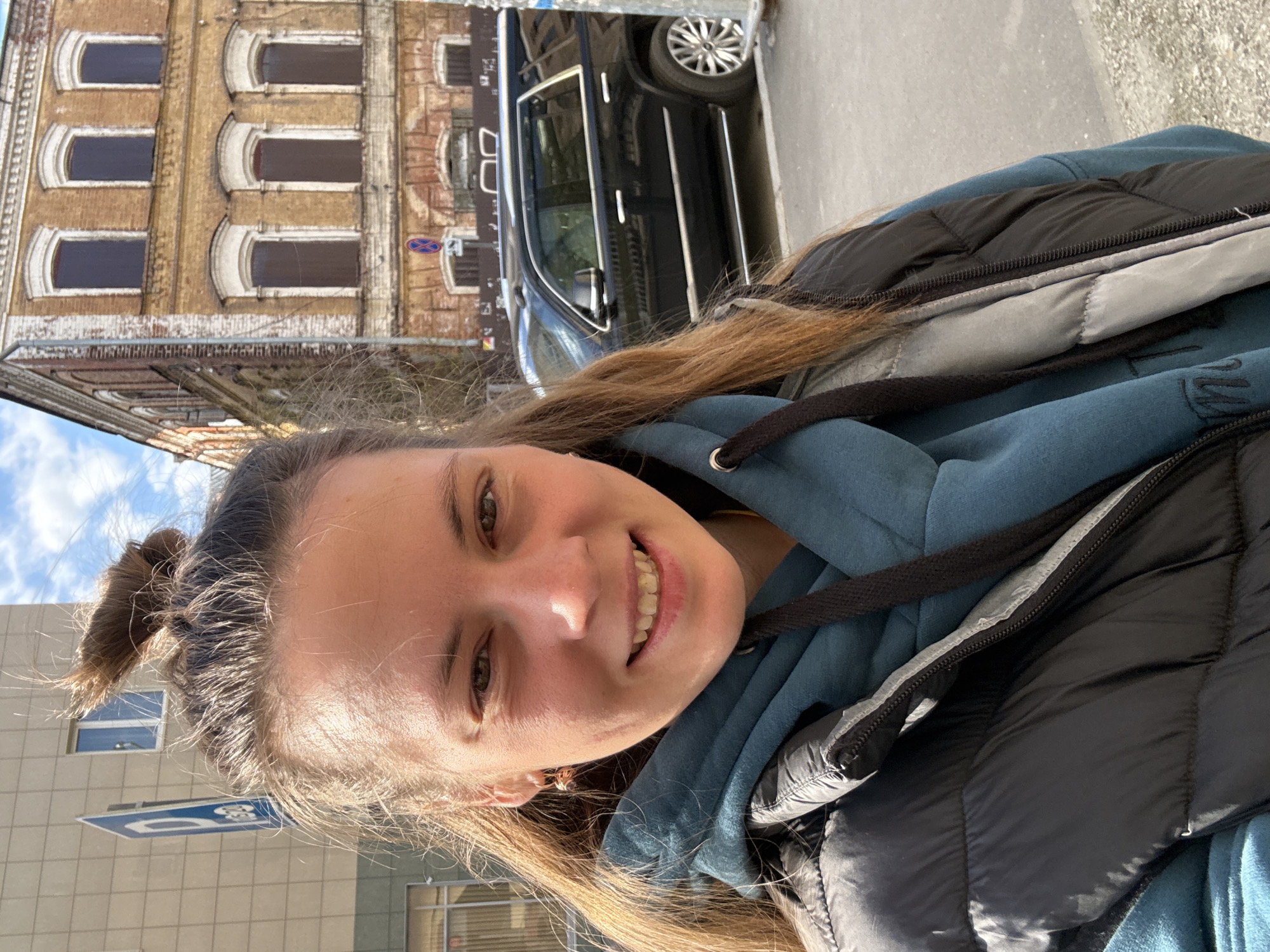 A woman stands on a street, a brown brick building with arched windows behind her, a black car parked nearby, and a blue parking sign visible to her left. She is smiling, facing towards the viewer, wearing a teal hoodie and a black puffer vest. The location is Kazan, Russia, based on the architectural style of the building and parking sign.