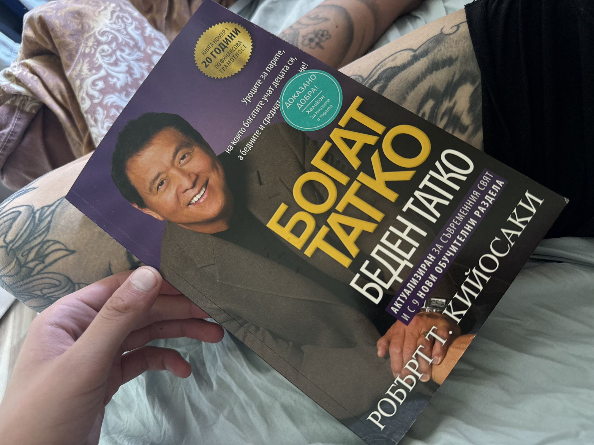 The image displays a close-up of a person holding a Bulgarian copy of Robert T. Kiyosaki's book "Rich Dad Poor Dad" (БОГАТ ТАТКО БЕДЕН ТАТКО). The scene is set indoors, likely on a bed or sofa, with patterned bedding in shades of purple, beige, and light blue visible in the background.
The book is held by a hand, revealing a thumb and fingers. Visible on the person's upper arm and shoulder are tattoos with abstract, flowing designs in dark ink. The activity taking place is either holding the book to read or preparing to read.
Notable details on the book cover include a smiling portrait of Robert Kiyosaki. The main title "БОГАТ ТАТКО БЕДЕН ТАТКО" is in large yellow letters on a black background, with the author's name "РОБЪРТ Т. КИЙОСАКИ" at the bottom. A prominent golden seal on the top left indicates "КНИГА НОМЕР 1 ПО ФИНАНСОВА ГРАМОТНОСТ 20 ГОДИНИ" (Book Number 1 for Financial Literacy 20 Years). Other text on the purple section of the cover reads "Уроците за парите, на които богатите учат децата си, а бедните и среднат не!" (The lessons about money that the rich teach their children, and the poor and middle class do not!). A circular green seal states "ДОКАЗАНО ДОБРА! Хеликон За книгите и хората" (Proven Good! Helikon For books and people). A subtitle below the main title specifies "АКТУАЛИЗИРАН ЗА СЪВРЕМЕННИЯ СВЯТ И С 9 НОВИ ОБУЧИТЕЛНИ РАЗДЕЛА" (Updated for the Modern World and with 9 New Educational Sections). The Bulgarian text on the book confirms the location context of Stara Zagora, Bulgaria.