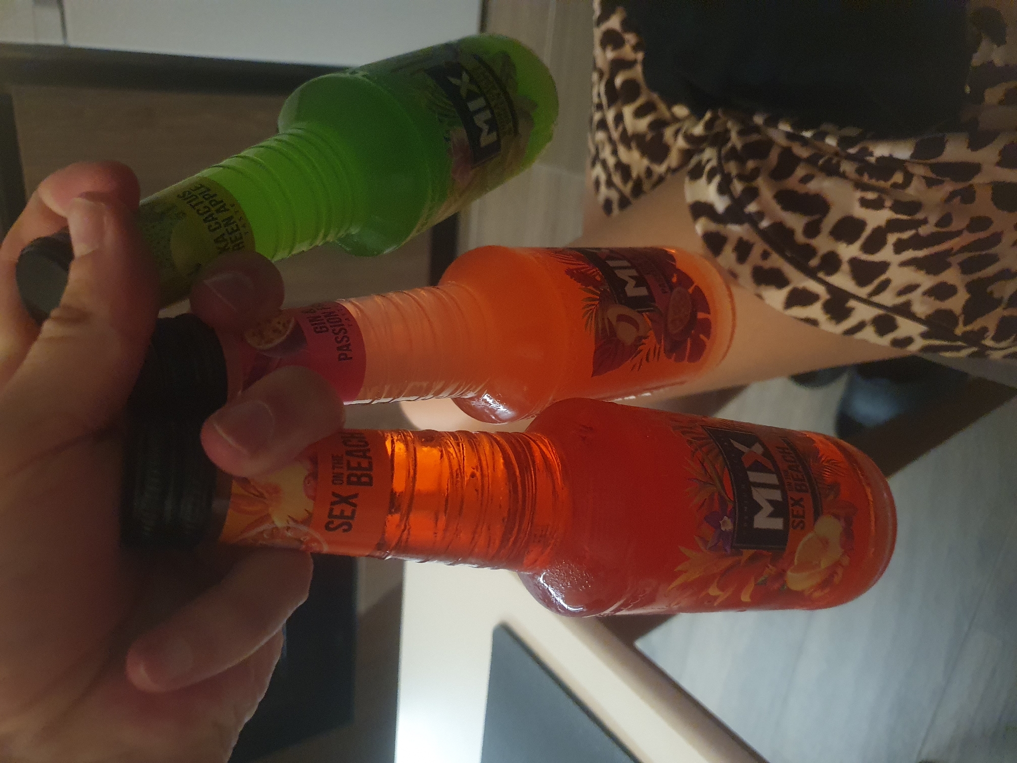 A person is holding three small bottles of "MIX" brand alcoholic beverages. The bottles are arranged in a stack, with the bottom bottle labeled "SEX ON THE BEACH," the middle bottle labeled "GIN PASSION FR," and the top bottle labeled "KA CACTUS GREEN APPLE." The person's hand is visible, gripping the bottles near the caps. The image appears to be taken indoors. The pattern on the person's clothing appears to be leopard print. There is a table visible in the background. The location in the city of Plovdiv, Bulgaria is not discernible from the image.