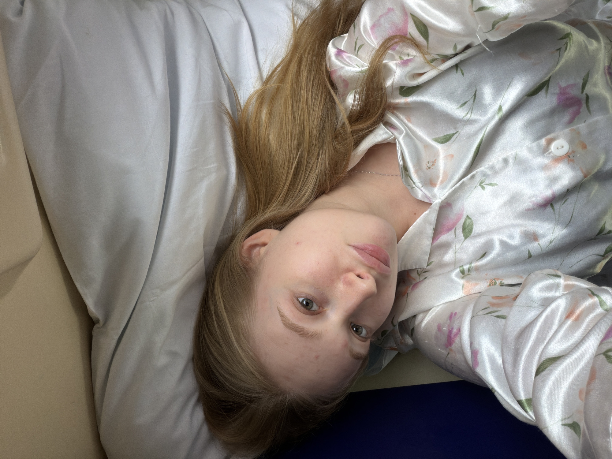 A woman is lying on her back on a bed. She has light blonde hair and fair skin. She is wearing a white satin pajama top with a floral pattern. The bed has a white sheet and a tan headboard. The location is unknown; there is no indication it is in Medvyodovskaya, Russia.