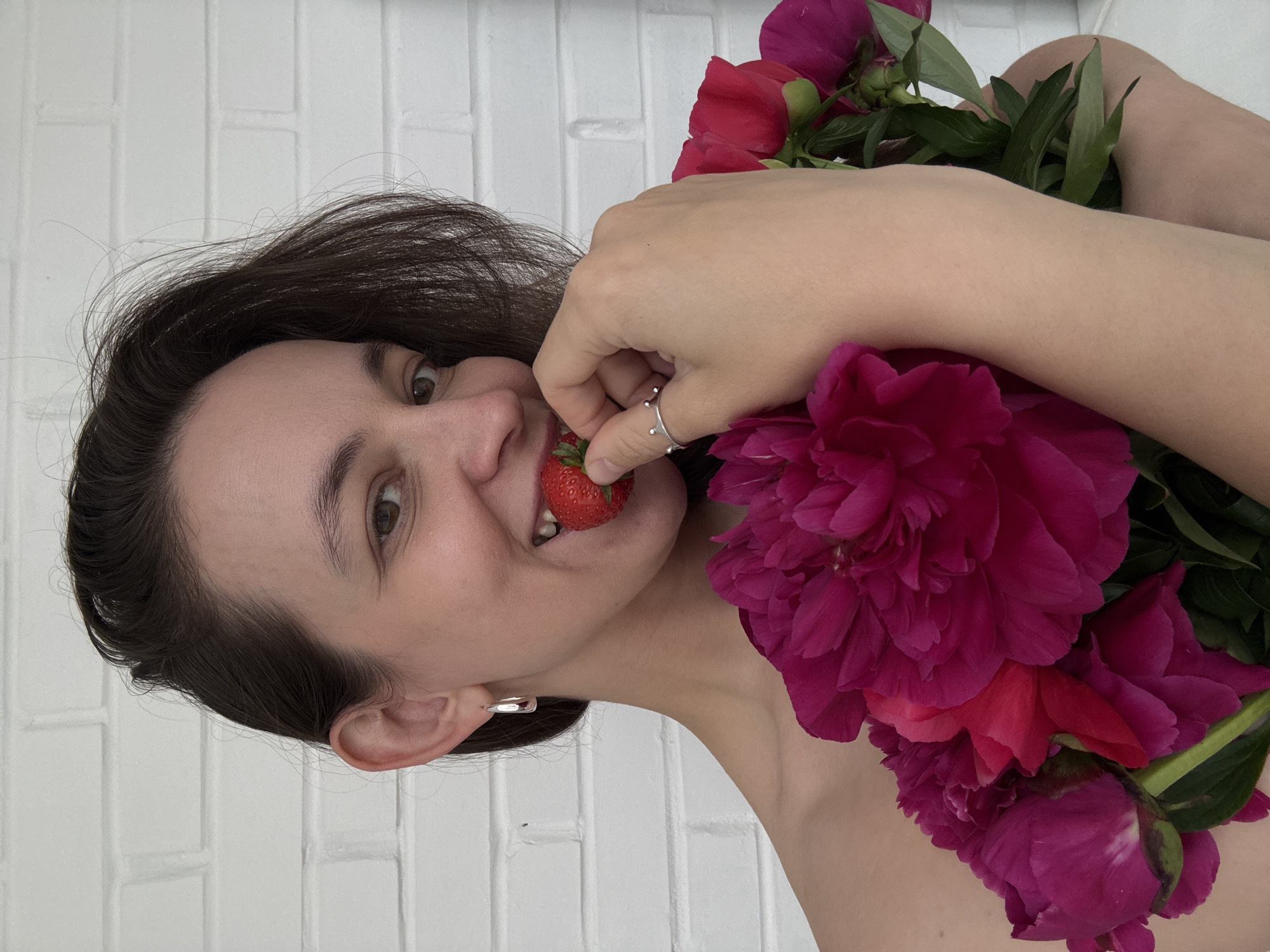 The scene, set in the city of Chaikovsky, Russia, features a woman smiling directly at the viewer. She has dark brown, shoulder-length hair and a light complexion, presented from the chest up with bare shoulders. Her left ear displays a silver-colored hoop earring. She holds a red strawberry between her teeth and her left hand, which is adorned with a silver-colored ring featuring a raised crown-like design on the index finger. Against her chest, she clutches a bouquet of multiple vibrant magenta and deep pink peonies. The background consists of a uniformly white-painted brick wall with clear horizontal mortar lines, illuminated by soft, even lighting.