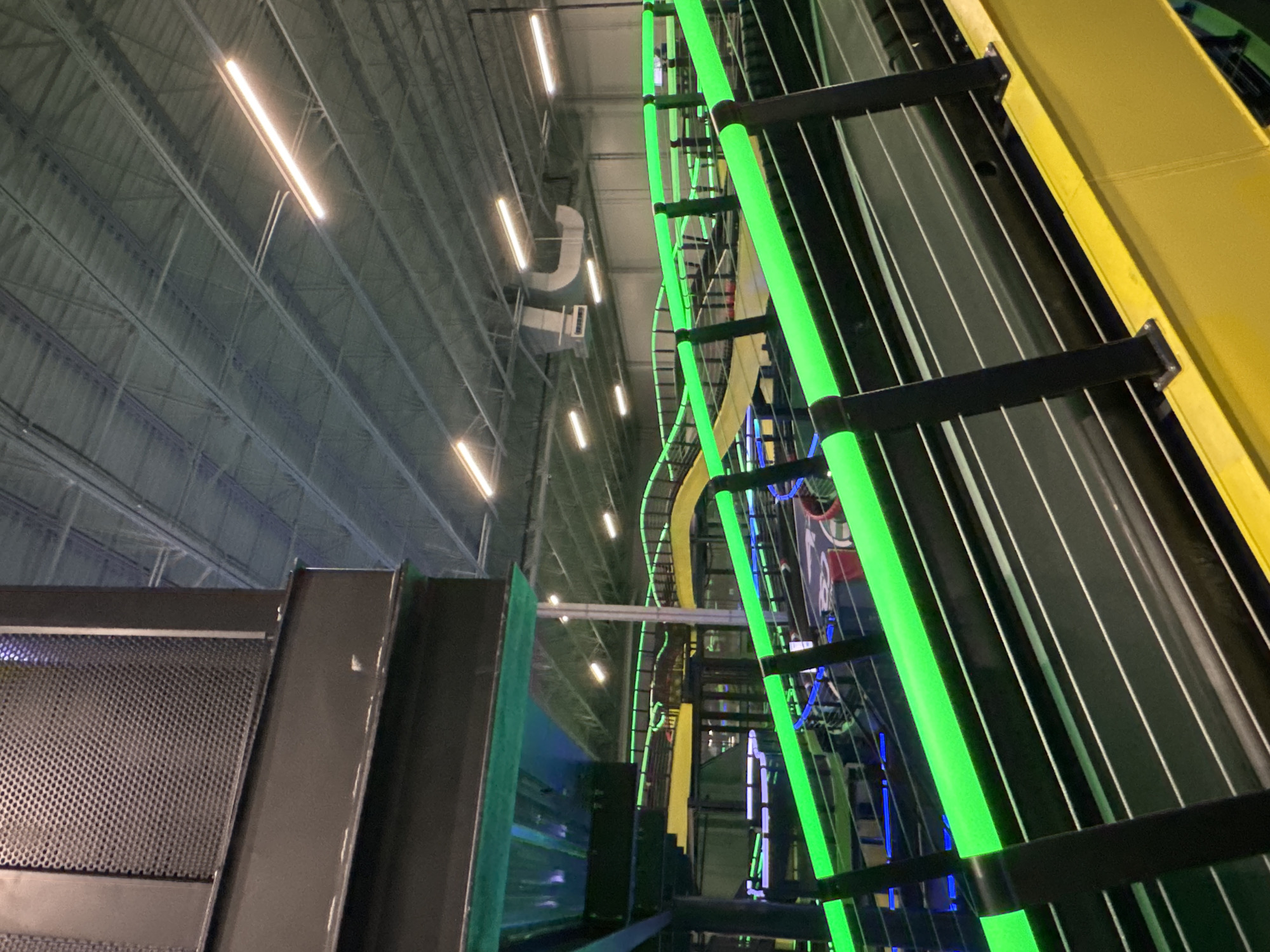The image shows an indoor recreational facility, likely a trampoline park or entertainment center, located in Schaumburg, Illinois, United States. The scene is characterized by a high ceiling with exposed metal beams and rows of fluorescent lights. The facility features various structures including what appear to be climbing walls, ramps, and elevated platforms. Some of these structures are illuminated with bright green neon lights. Metal safety netting and framing are prominent throughout the scene. A section of a gray metal structure with perforated paneling is visible in the foreground.